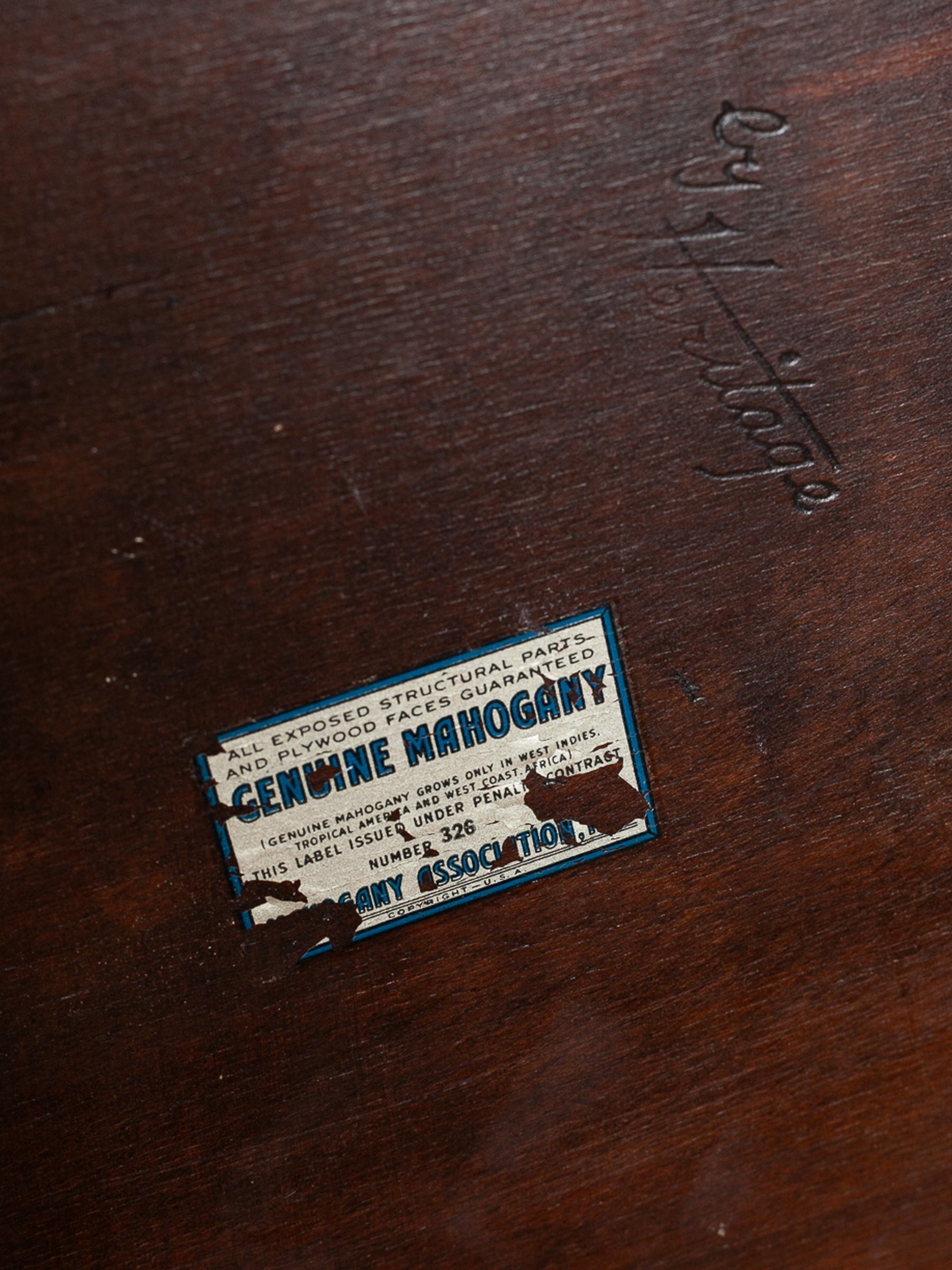 A close-up of the underside of a wooden table with a small worn paper label reading 'Genuine Mahogany' and a dark embossed script logo "by Heritage" above it.