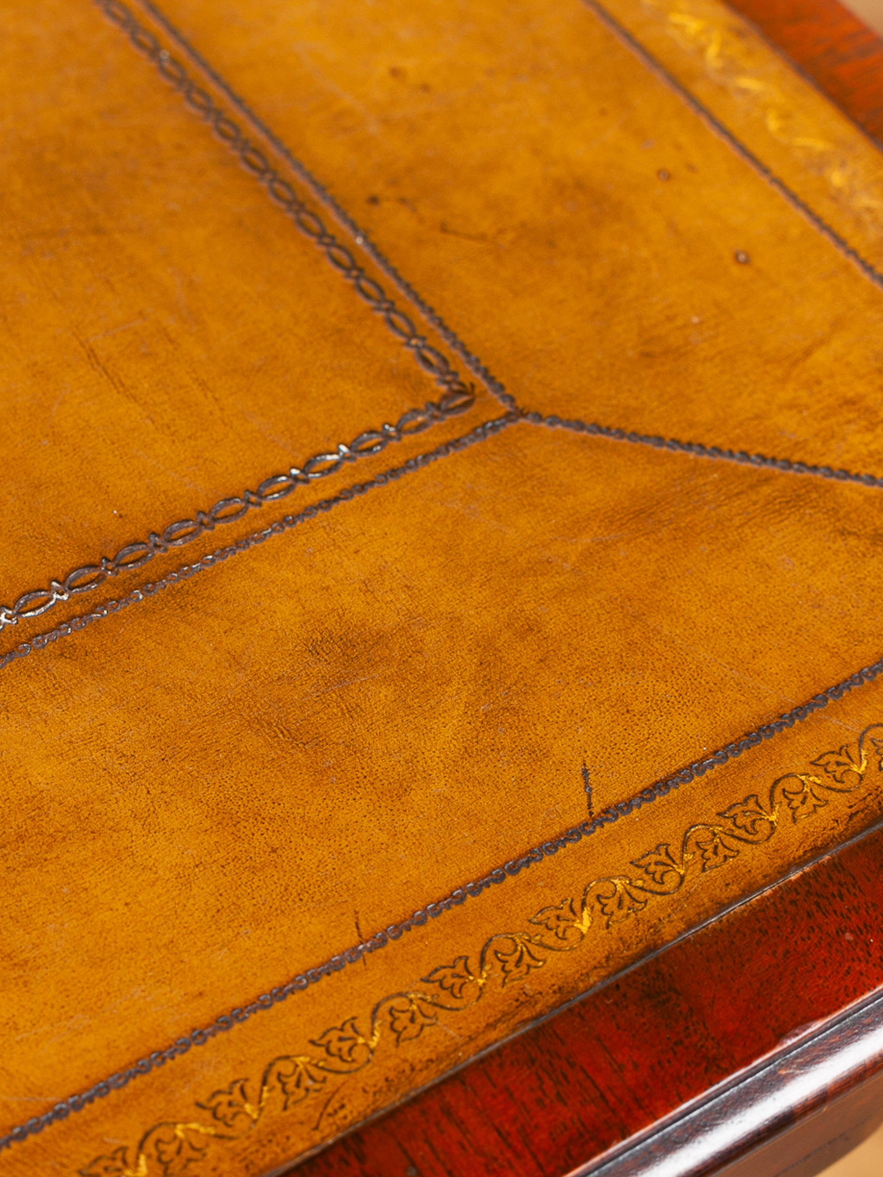 Close-up of an antique leather top table warm olive leather with stitched geometric panel lines and embossed decorative border details, resting in a dark wood surface.