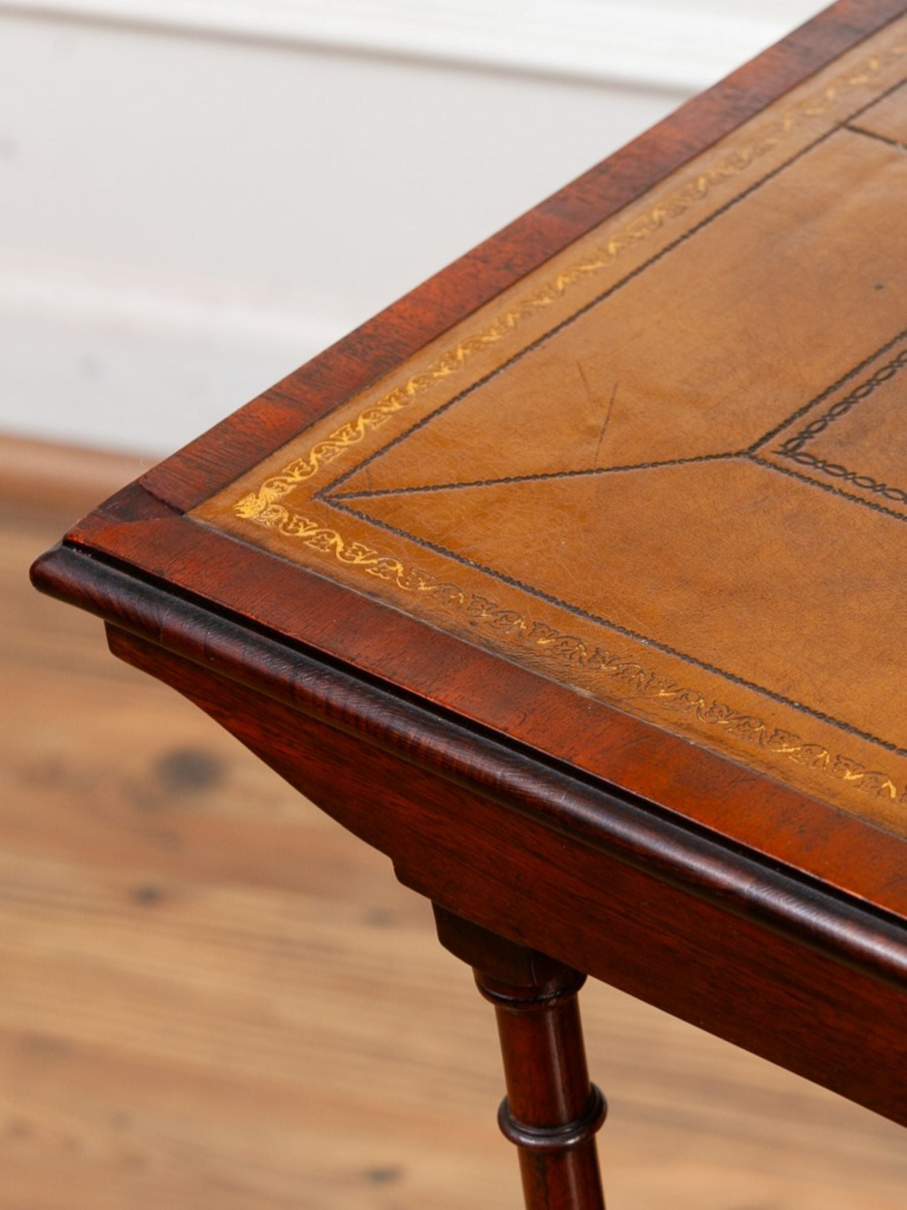 Close-up of a wooden table corner showing a warm olive leather inlay with decorative border detailing and a dark reddish-brown wood frame and leg.