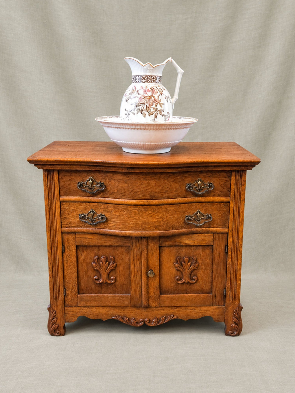 A small vintage-style wooden side table or Washstand with carved details and dark metal drawer pulls, shown against a neutral draped fabric backdrop. A white ceramic pitcher and matching bowl sit on top.