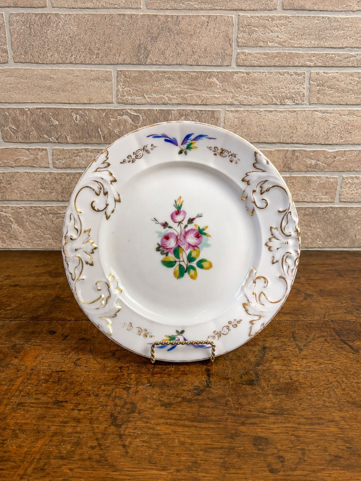 A vintage white porcelain plate with gold scrollwork and a central pink rose motif, displayed on a wooden surface against a brick wall background.