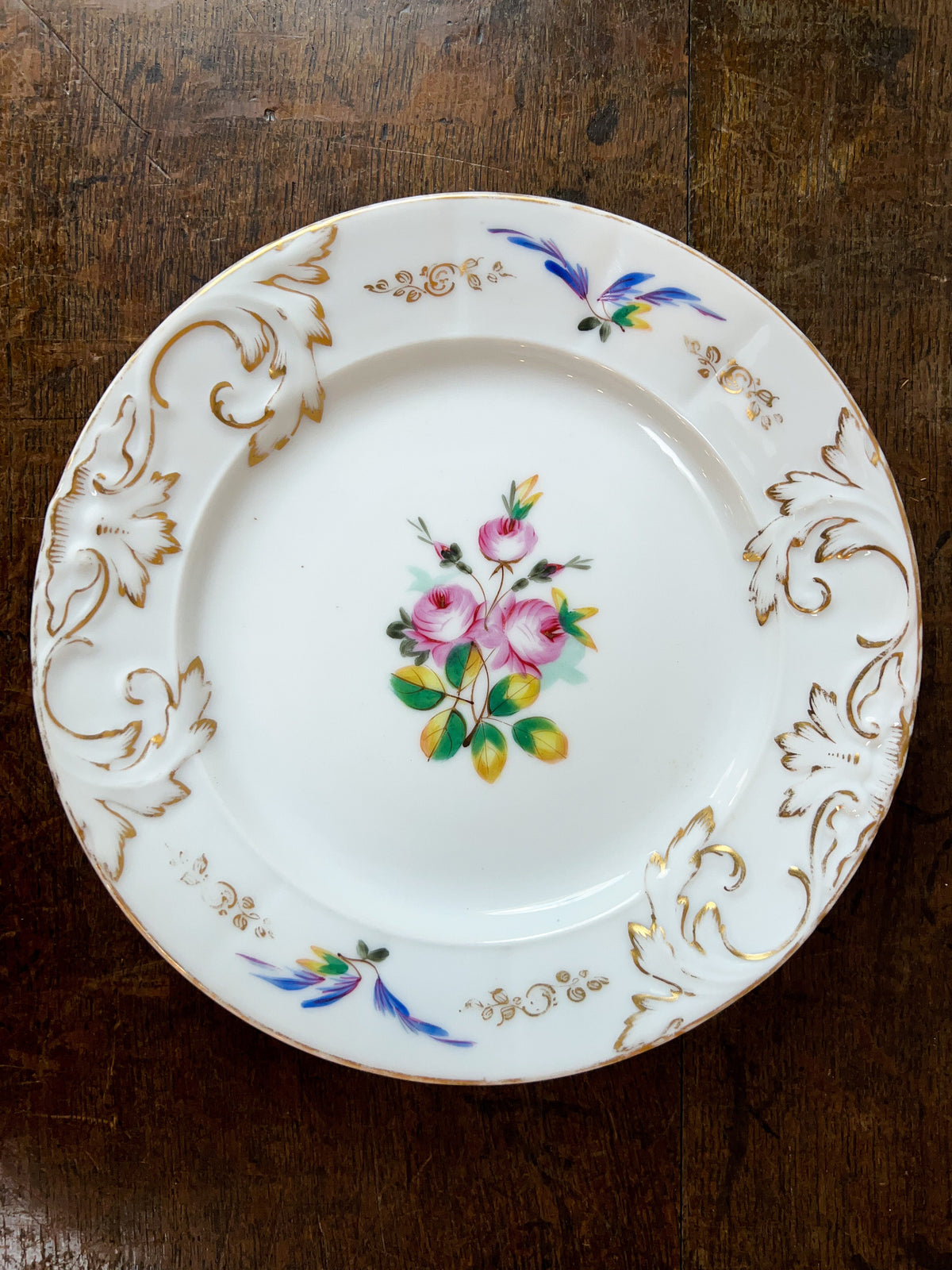 A vintage white porcelain plate with gold leaf detailing, featuring a central pink rose floral motif and blue and gold scrollwork, displayed on a dark wooden surface.