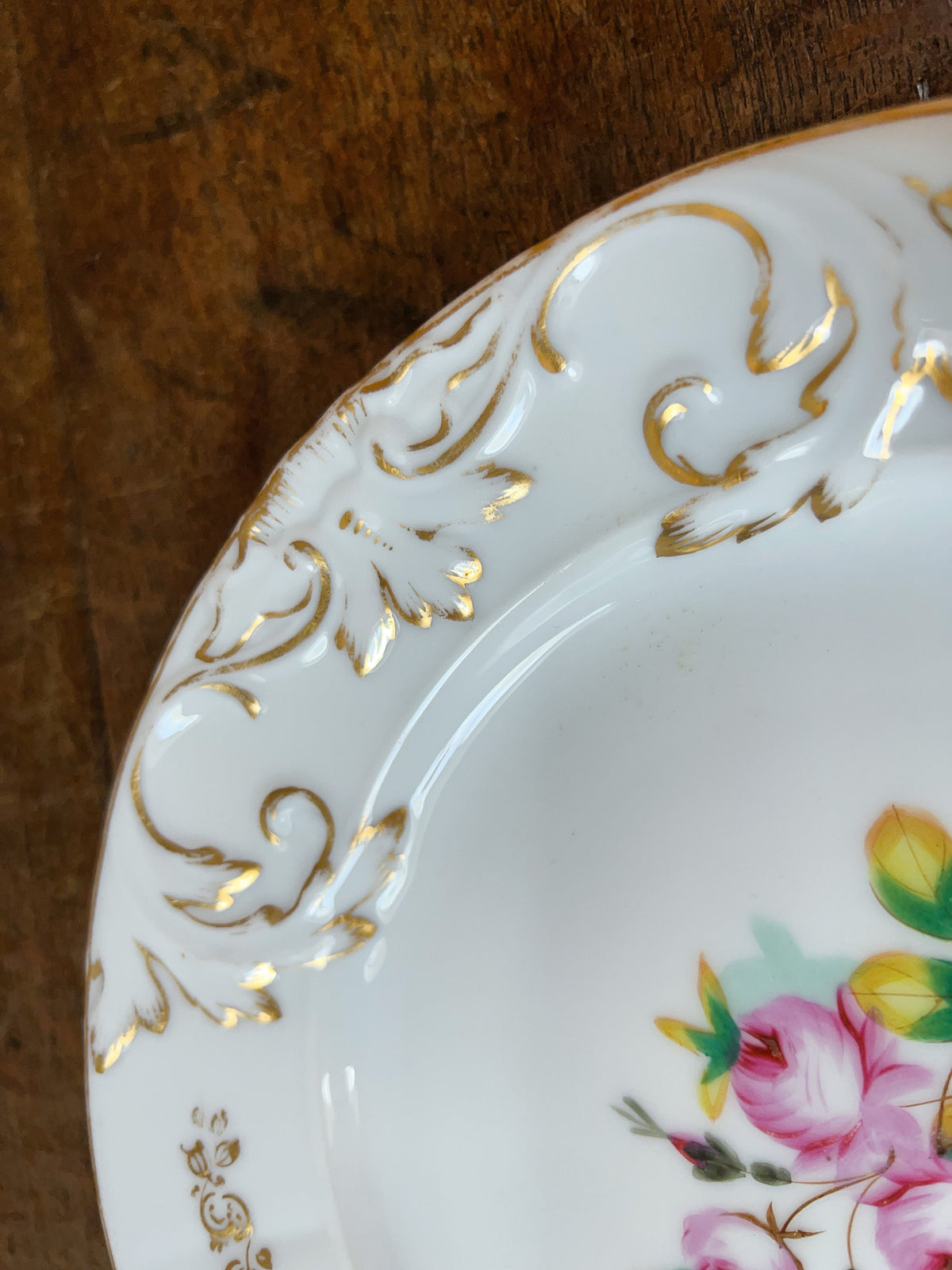 A close-up of a white porcelain plate with ornate gold embossed scrollwork on the rim and a partial floral print in pink, green, and yellow, set against a dark wooden background.
