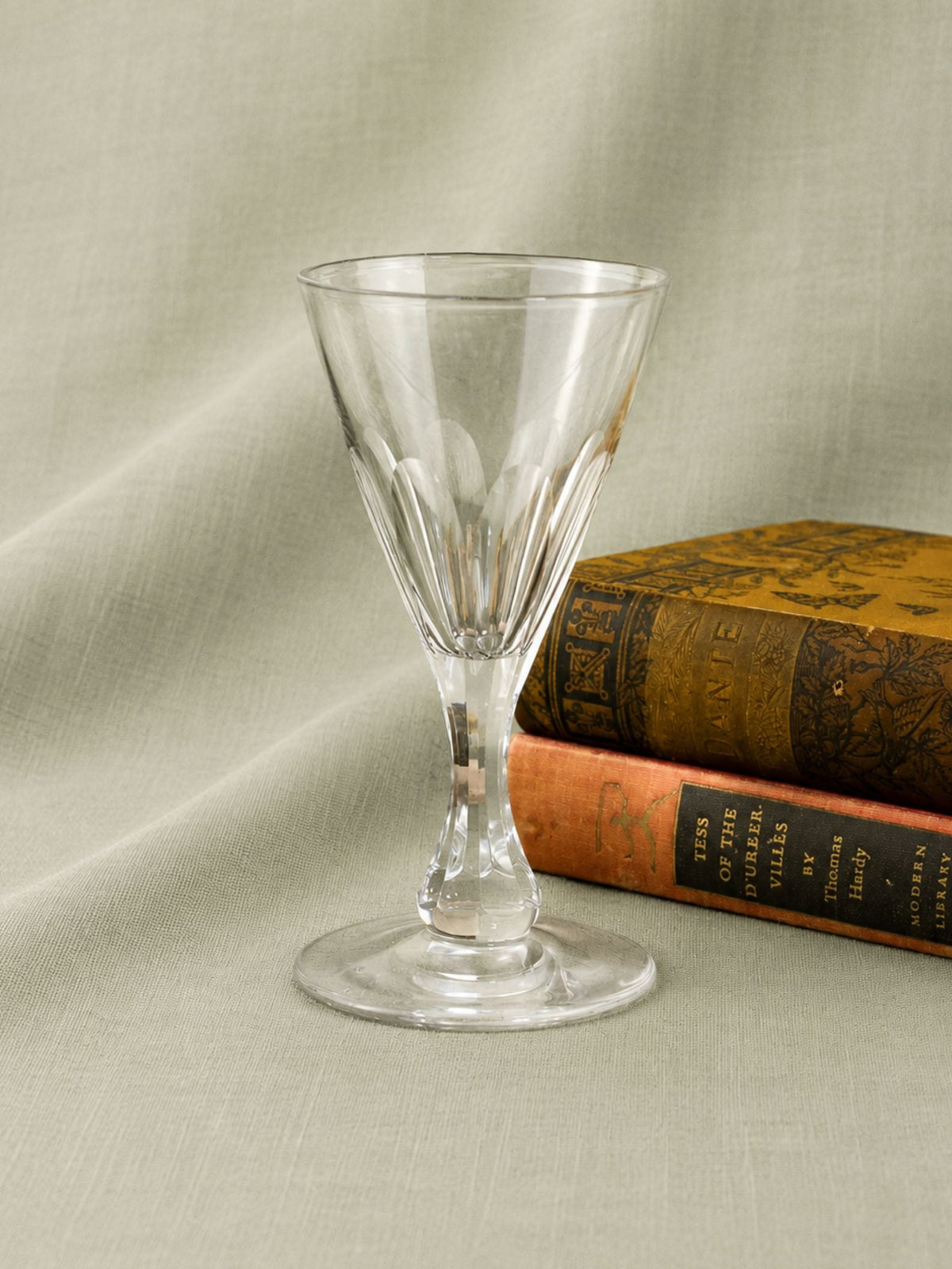 A clear cut-glass goblet or tumbler with a flared rim and pedestal base, photographed on draped light beige fabric with two vintage books behind it.