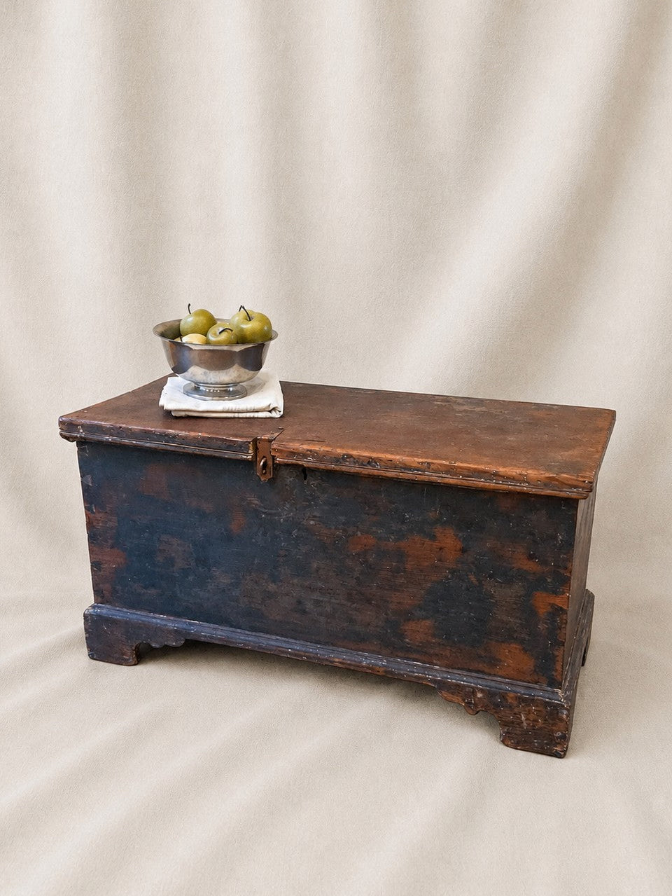 A rustic dark brown wooden chest with a weathered black-painted front, styled with a small silver bowl of pears on top, set against a draped beige fabric backdrop.