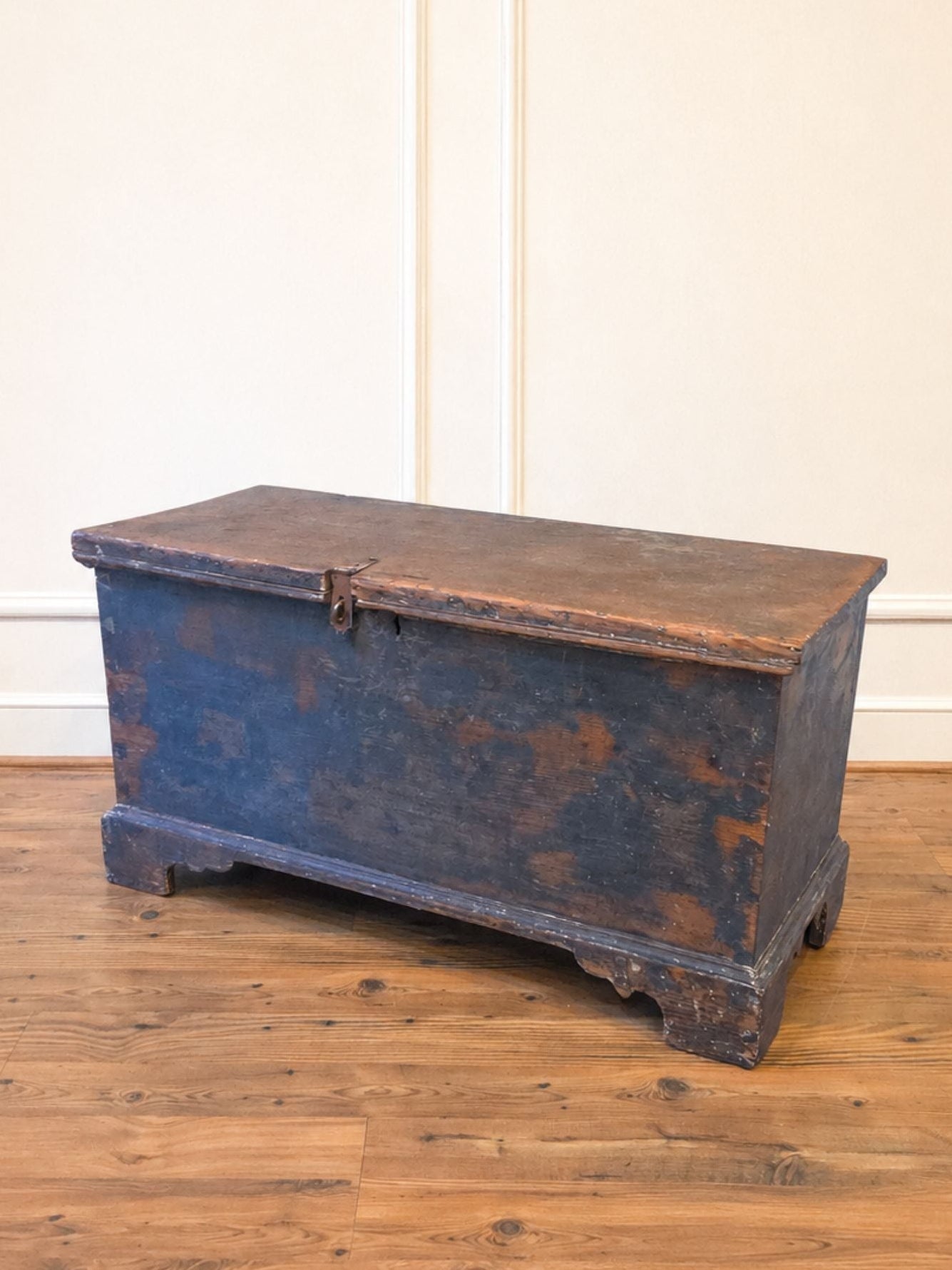 A rustic dark blue wooden chest with a weathered brown top, placed on a light brown wooden floor against a cream paneled wall.