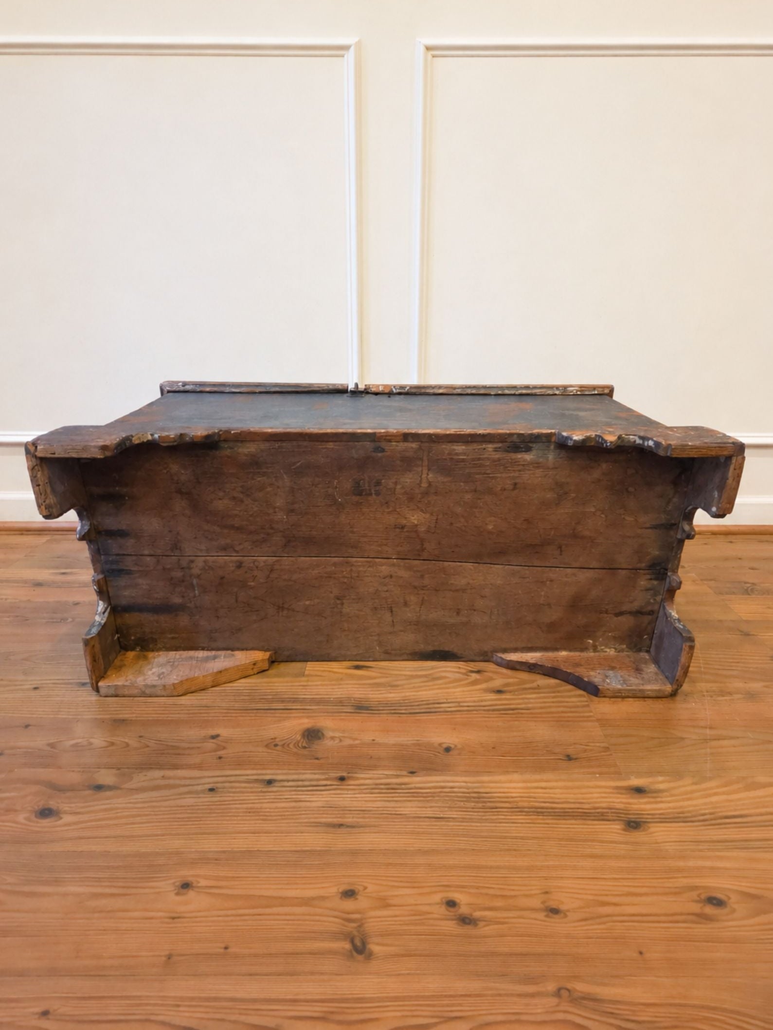 The underside of a rustic dark brown wooden chest or bench with a weathered finish, placed on a polished wooden floor against a white paneled wall.