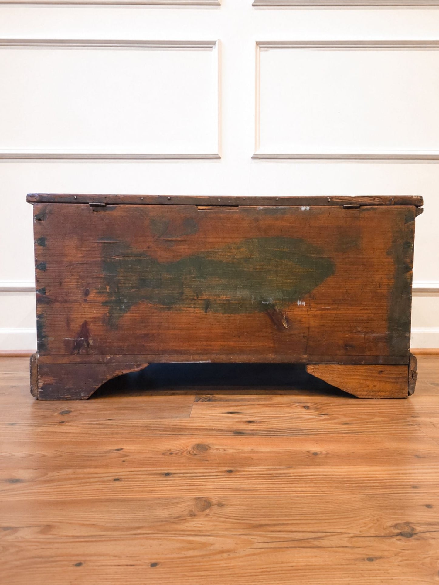 A rustic, vintage wooden chest with a dark brown finish and weathered greenish paint on the front, set on a light wood floor against a white paneled wall.