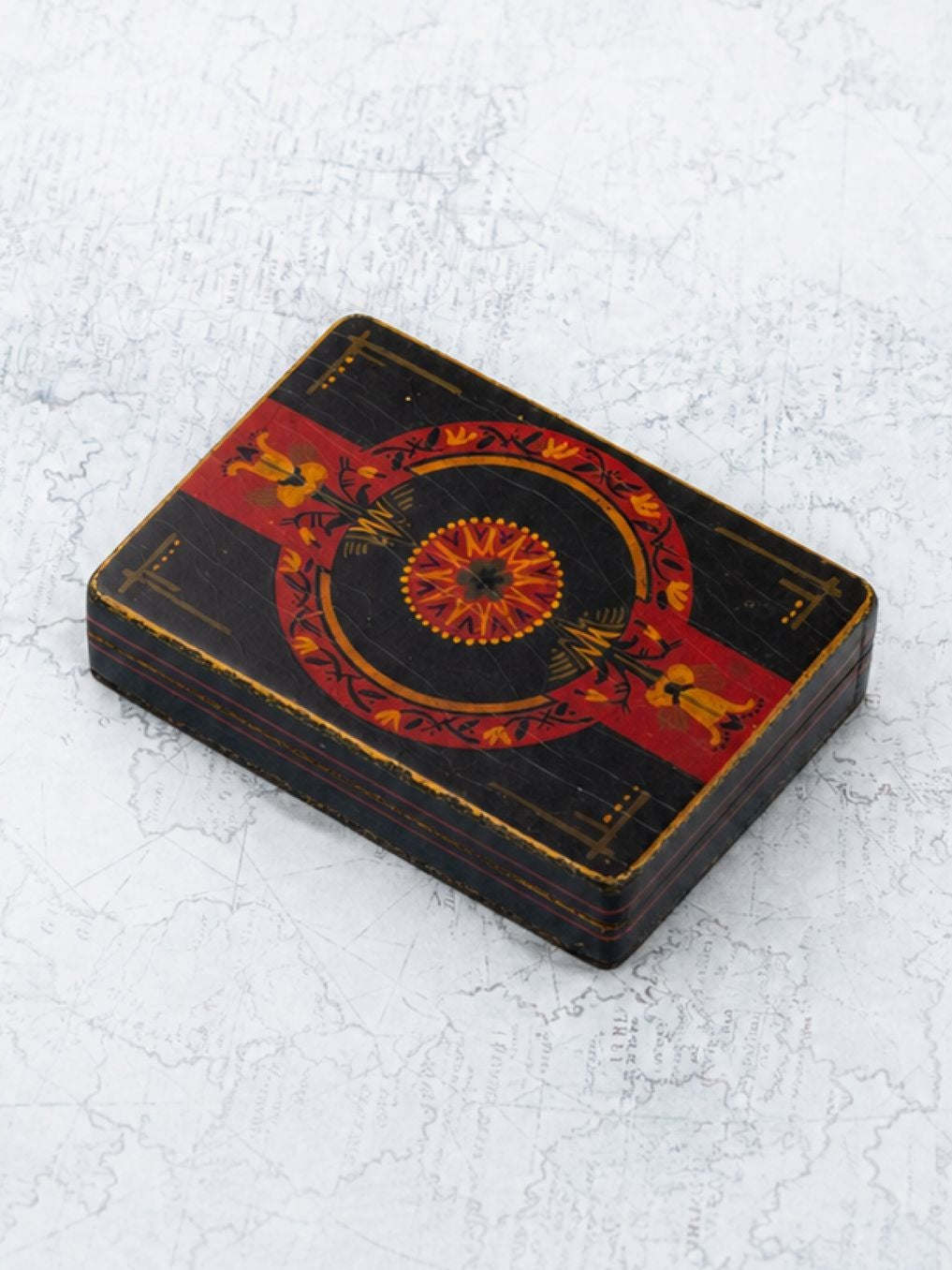 A small rectangular black decorative box with red and gold ornamental patterns, photographed on a light textured white background.