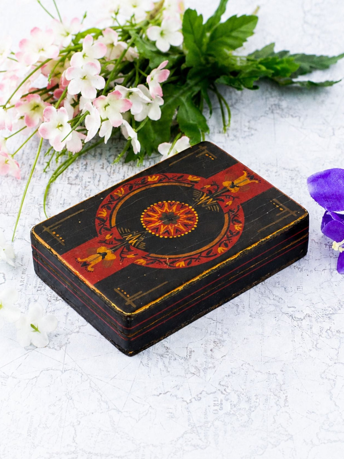 A rectangular black decorative box with red and gold floral patterns sits on a light textured surface, styled with small white and pink flowers on the left and a purple flower on the right.