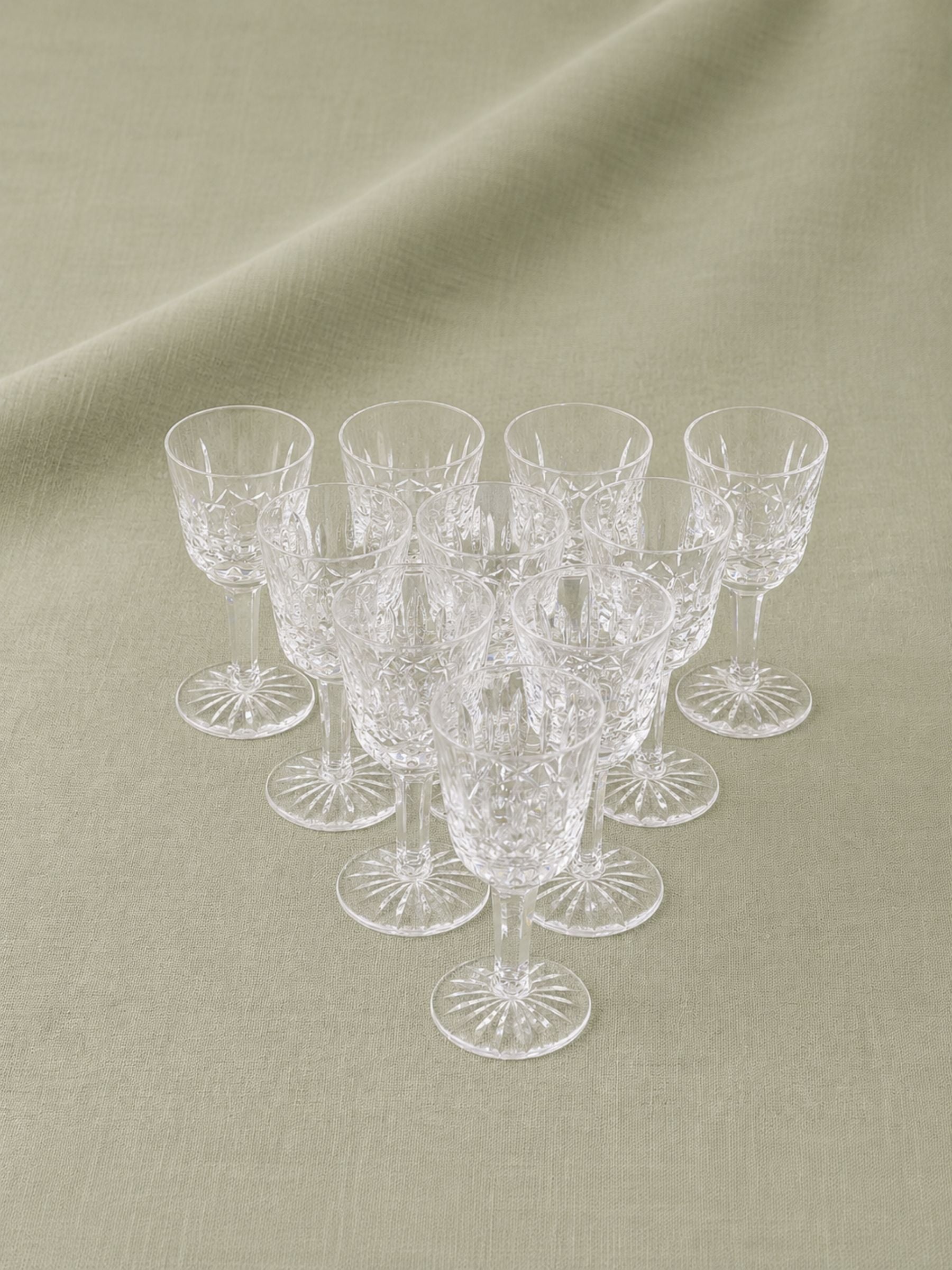 A set of clear cut-crystal aperitif glasses arranged in a cluster on a draped beige fabric background.