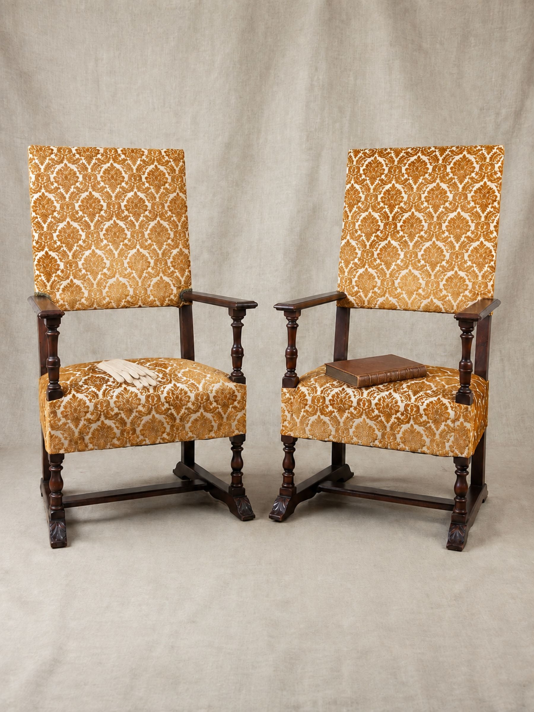 A pair of oversized wooden armchairs with dark brown turned wood frames and high backs, upholstered in a warm ochre yellow and beige damask patterned fabric, set against a neutral beige backdrop.
