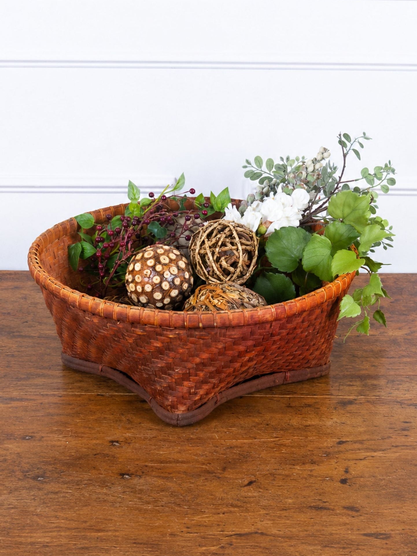 A rustic woven wicker basket filled with green foliage, white flowers, and decorative pinecones, set on a wooden table against a white paneled wall.