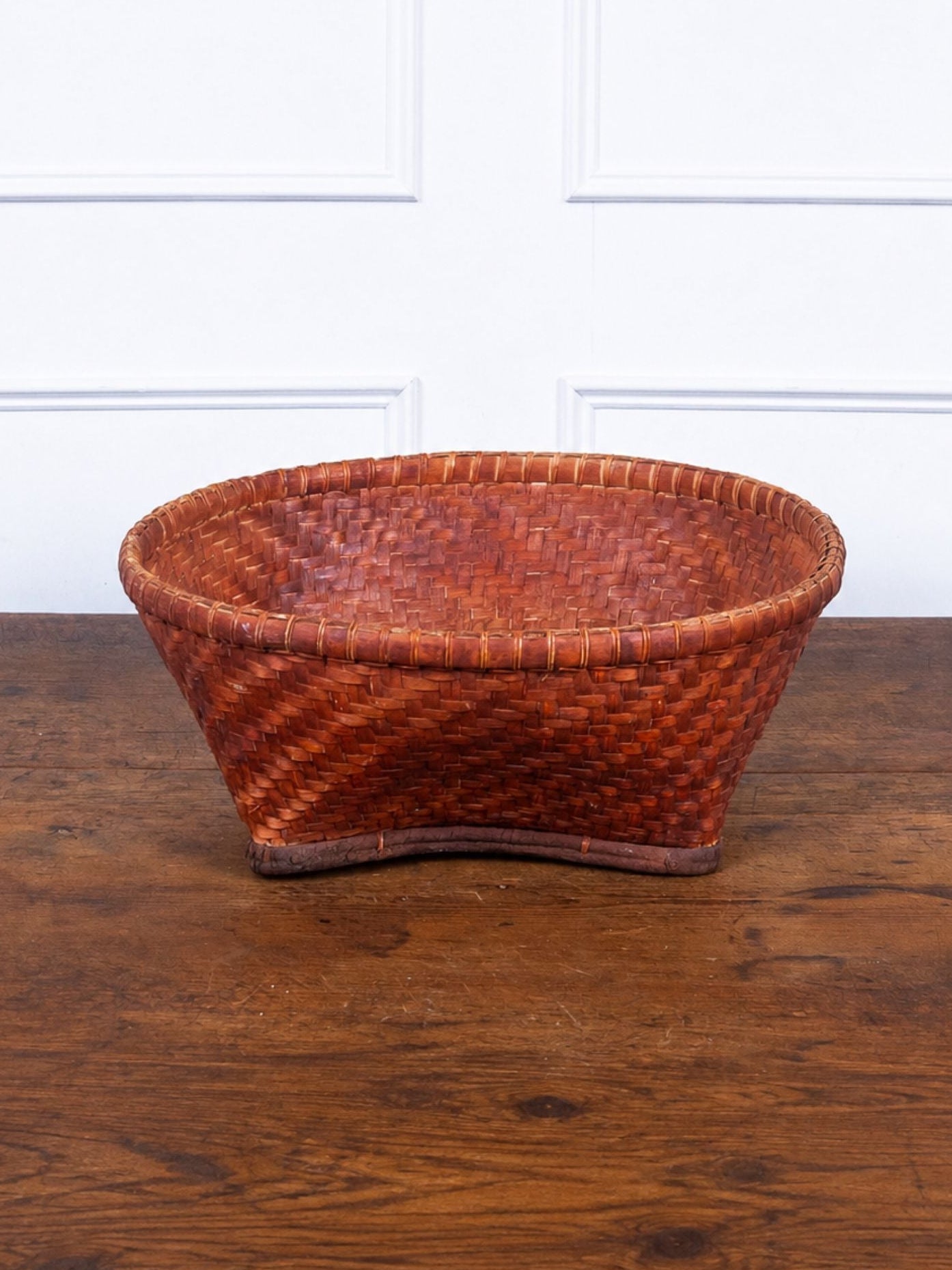 A round, hand-woven basket with a warm reddish-brown hue, displayed on a rustic wooden table against a white paneled wall.