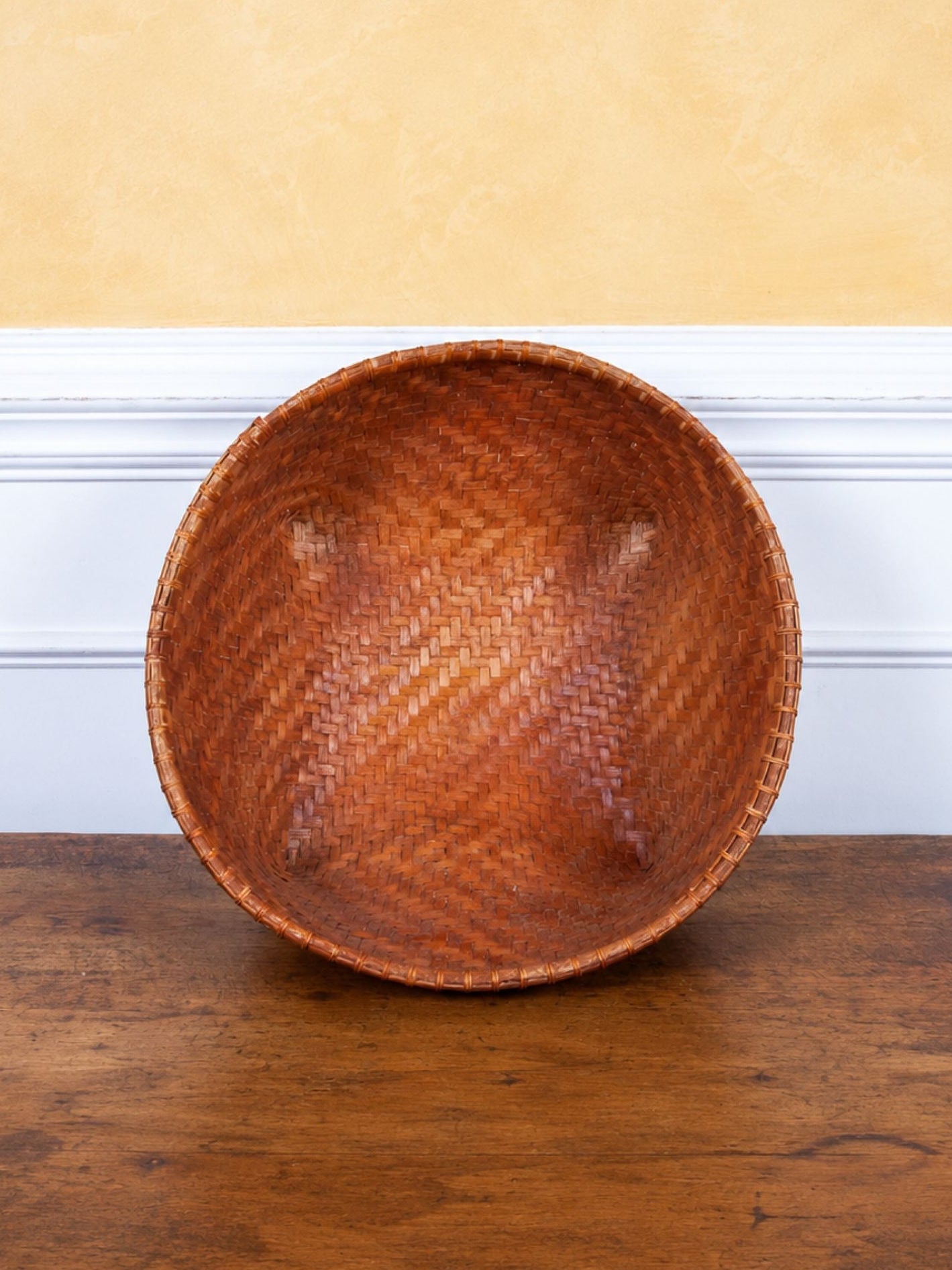 A round woven wooden bowl with a warm brown finish, shown standing on a dark wood tabletop against a white baseboard and textured yellow wall.