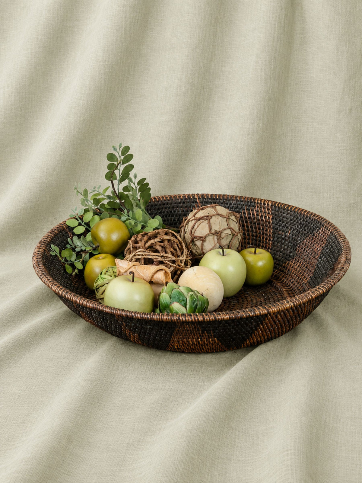 A dark brown woven oval tray filled with decorative fruits, greenery, and textured accents, set against a draped beige fabric backdrop.
