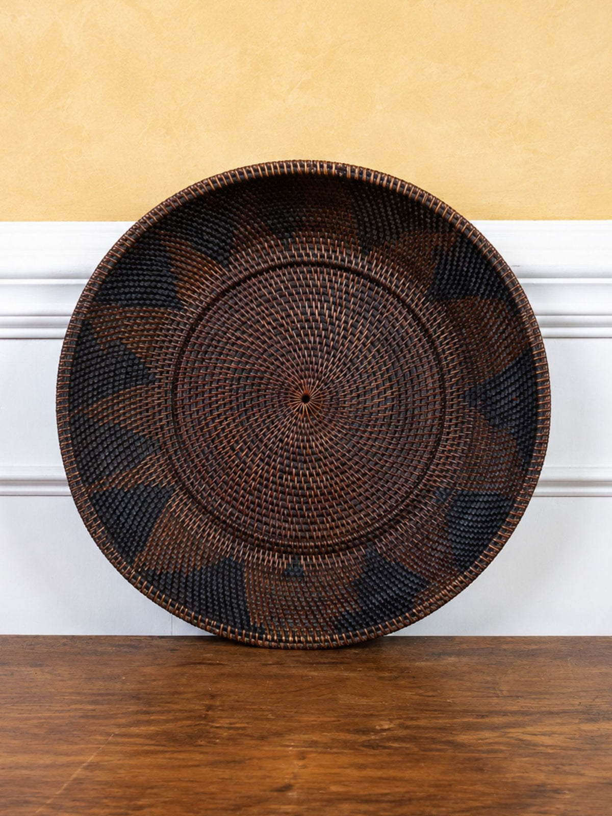 A round woven basket with a dark brown and black geometric pattern, leaning against a white chair rail on a wooden floor in front of a textured yellow wall.
