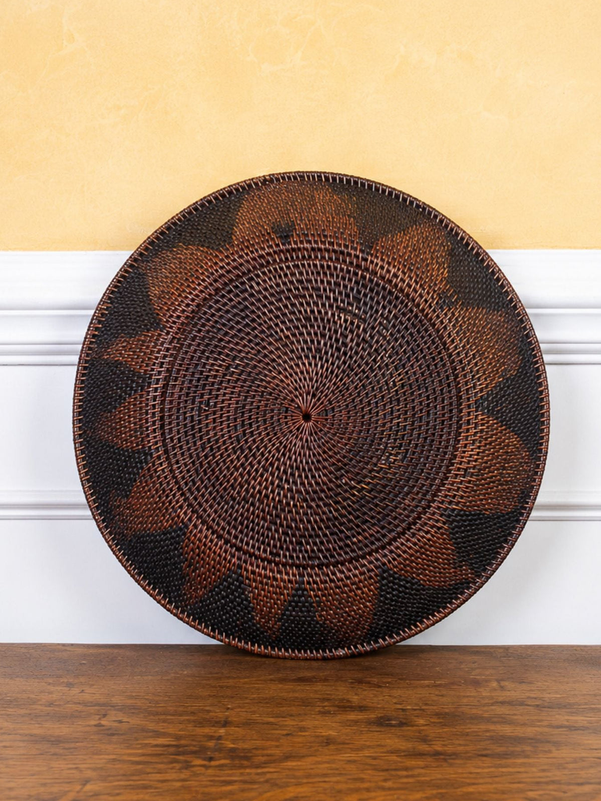 A round woven basket with a geometric starburst pattern in dark brown and black, leaning against a white chair rail on a textured yellow wall above a wooden table.