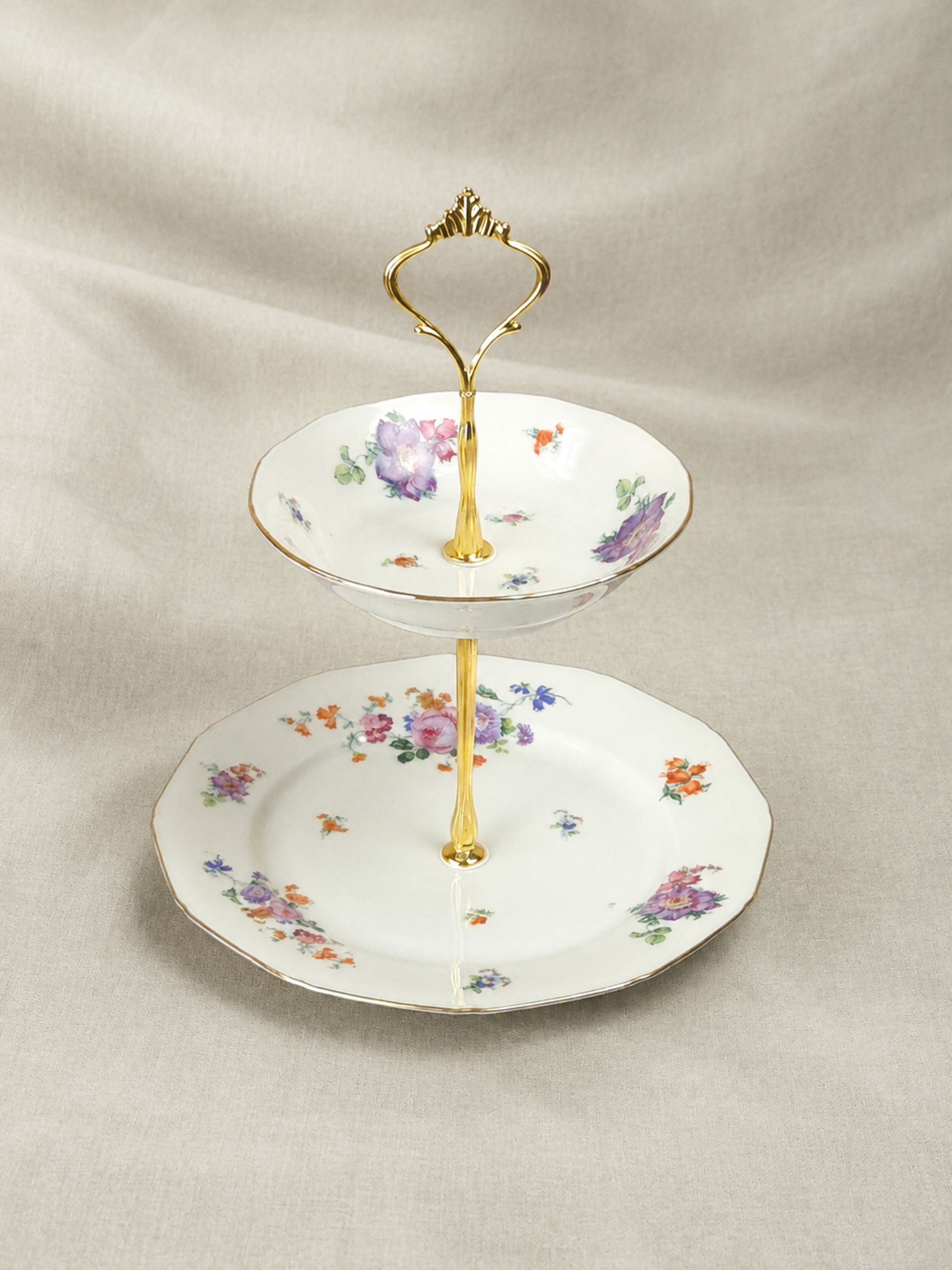Two-tier porcelain cake stand with colorful floral decoration, gilded rims and ornate gold-tone handle against a soft sage linen backdrop.
