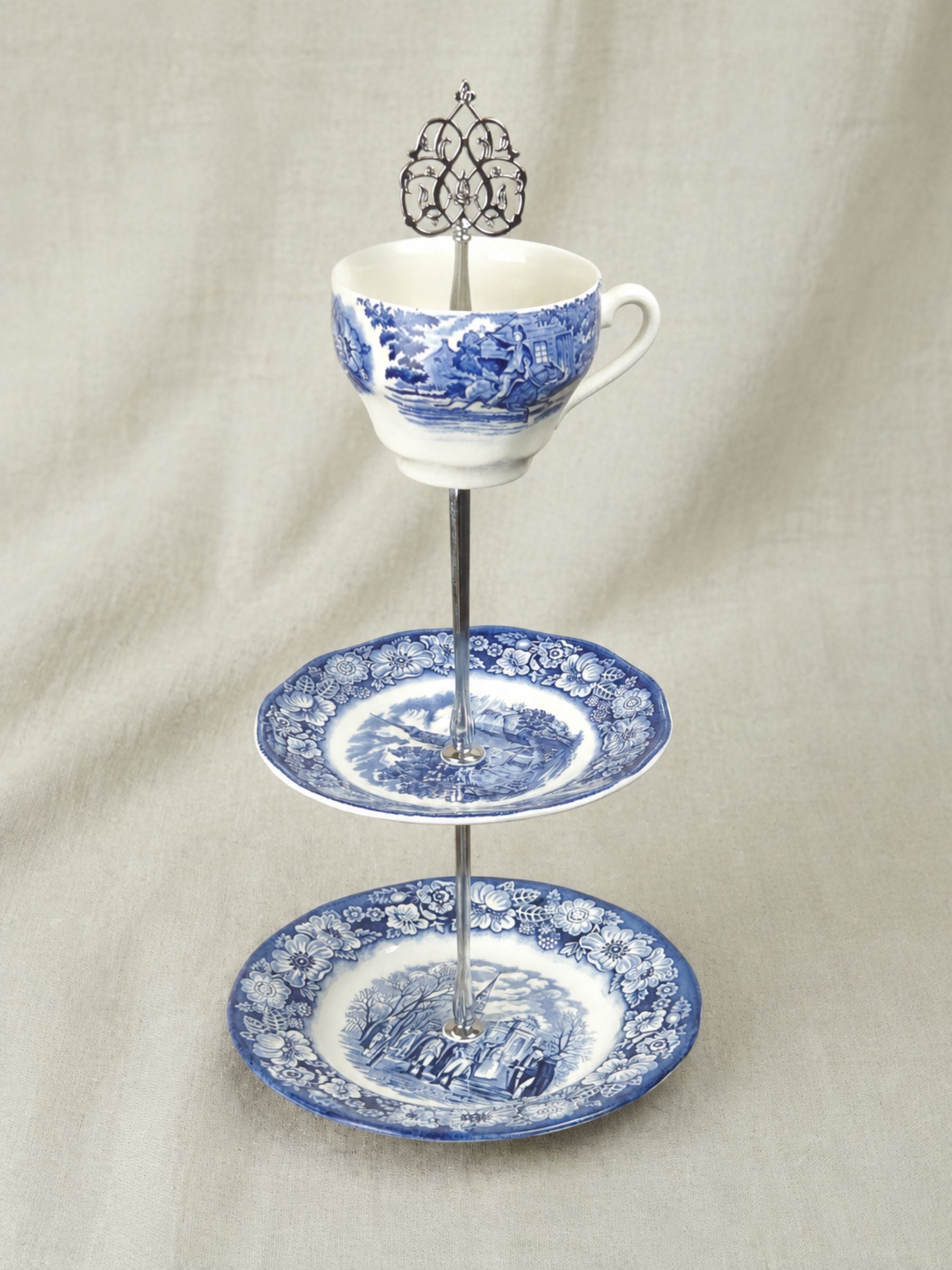A three-tier ceramic serving stand made from a teacup, saucer, and plate, decorated in a blue and white landscape pattern, set against a neutral draped fabric background.