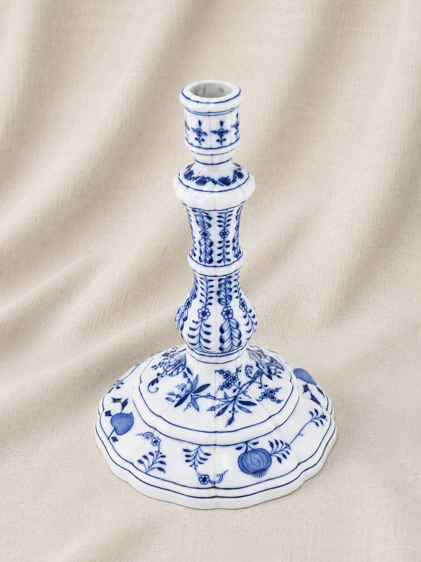 A blue and white porcelain candlestick holder with intricate floral and fruit motifs, set against a draped beige fabric background.