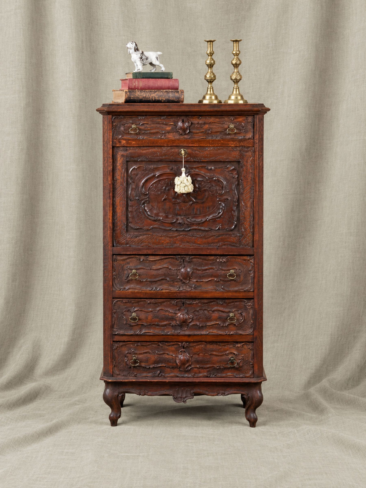 An antique carved wooden chest of drawers with a secretary drop front top and three drawers, ornate drawer pulls, and a hanging tag, shown on a draped beige fabric background with decorative books, a small figurine, and brass candlesticks on top.