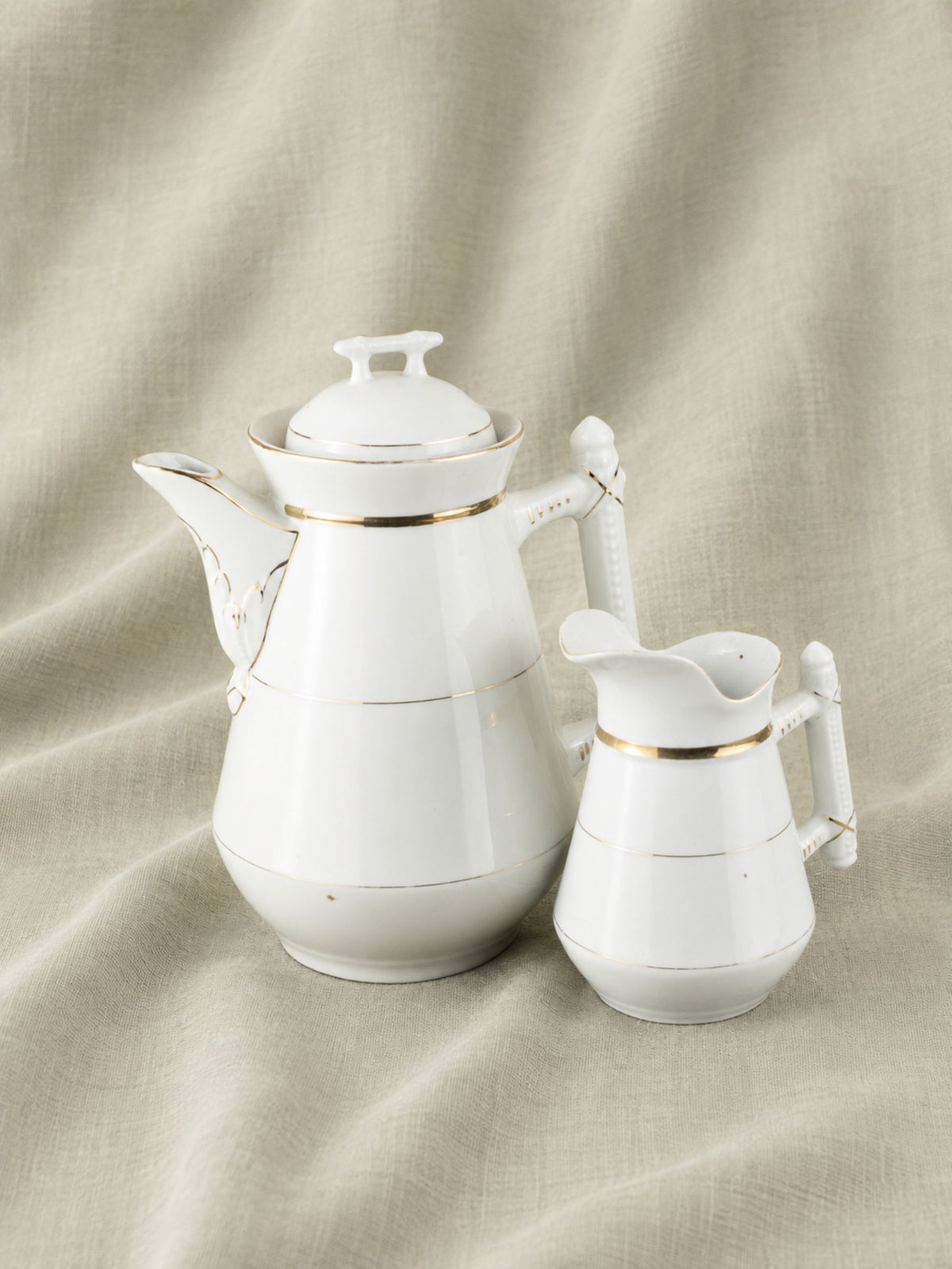 A white porcelain coffee pot and matching creamer pitcher with thin gold trim, set against a draped beige linen fabric background.