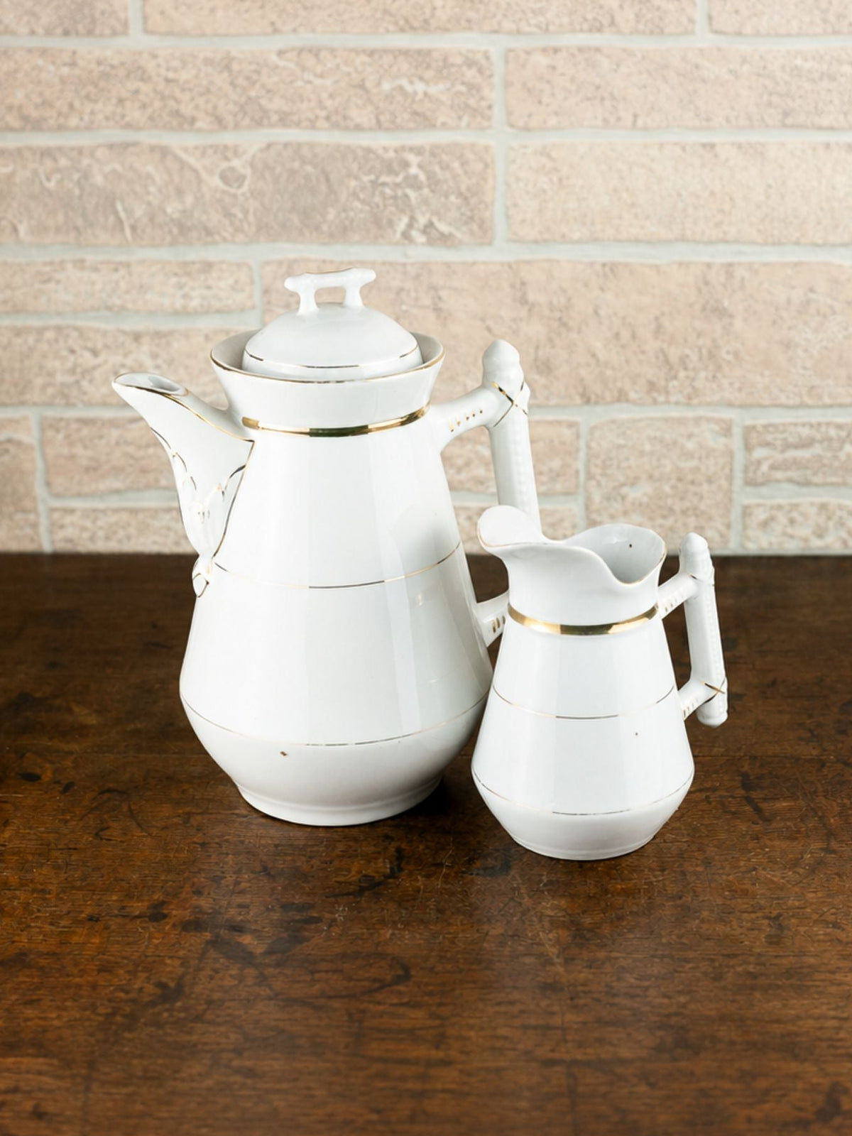 A white ceramic coffee pot and matching creamer pitcher with thin gold trim, set on a dark wooden table against a light beige brick wall background.
