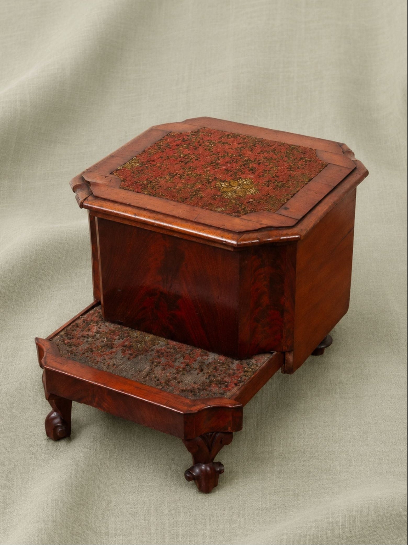 A vintage octagonal wooden commode and steps with a reddish-brown wood finish and a floral-patterned fabric top, set against a draped beige fabric background.