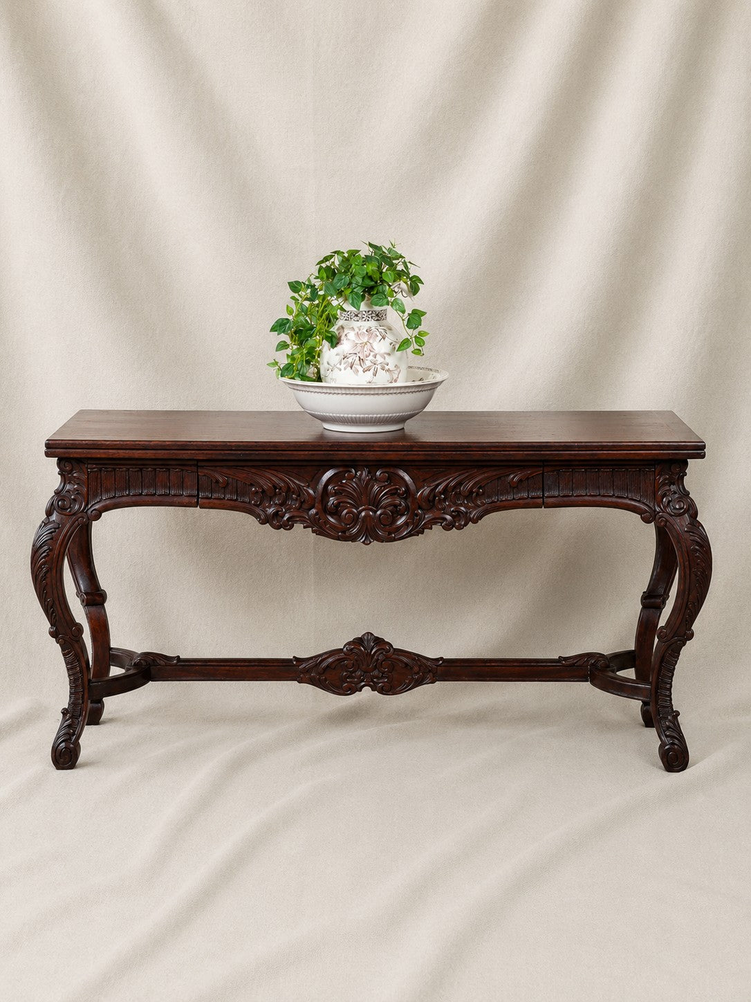 An ornate dark brown carved wooden fold out console table with cabriole legs, styled with a white bowl and green plant on top, set against a draped beige fabric backdrop.