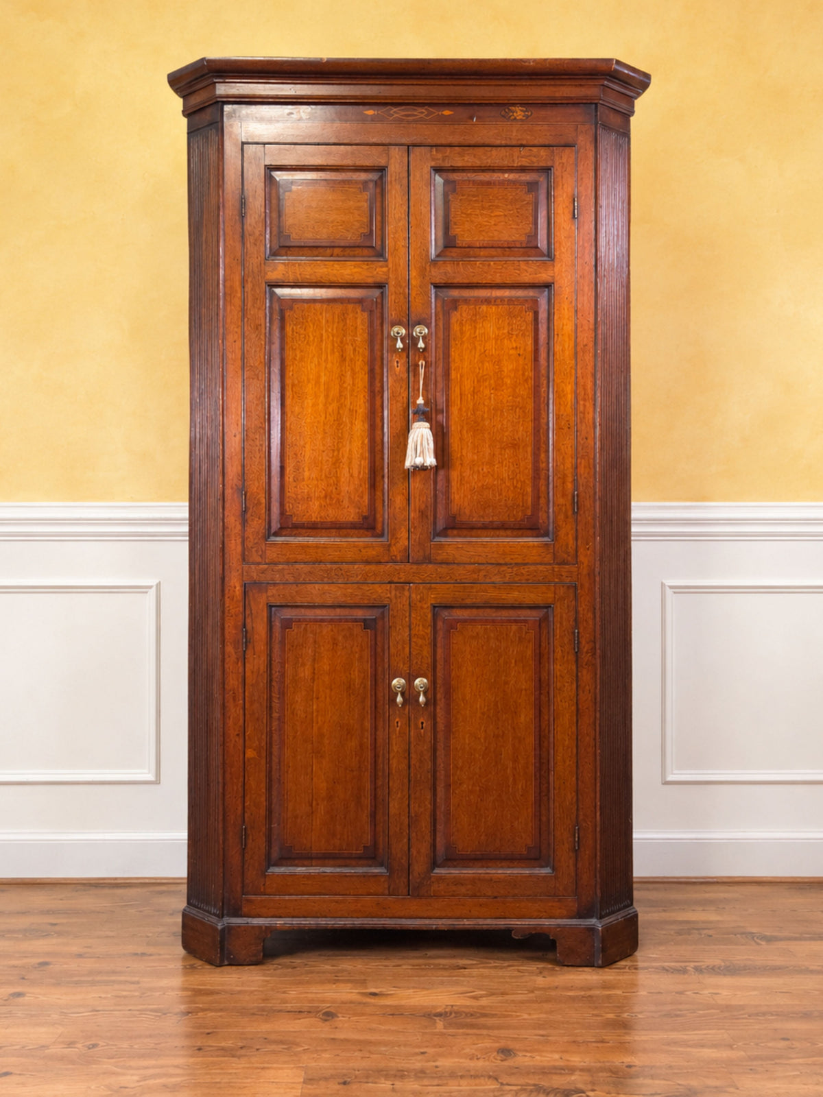 A tall, antique-style wooden corner cabinet with two paneled doors and brass hardware, set against a yellow wall with white wainscoting and a hardwood floor.