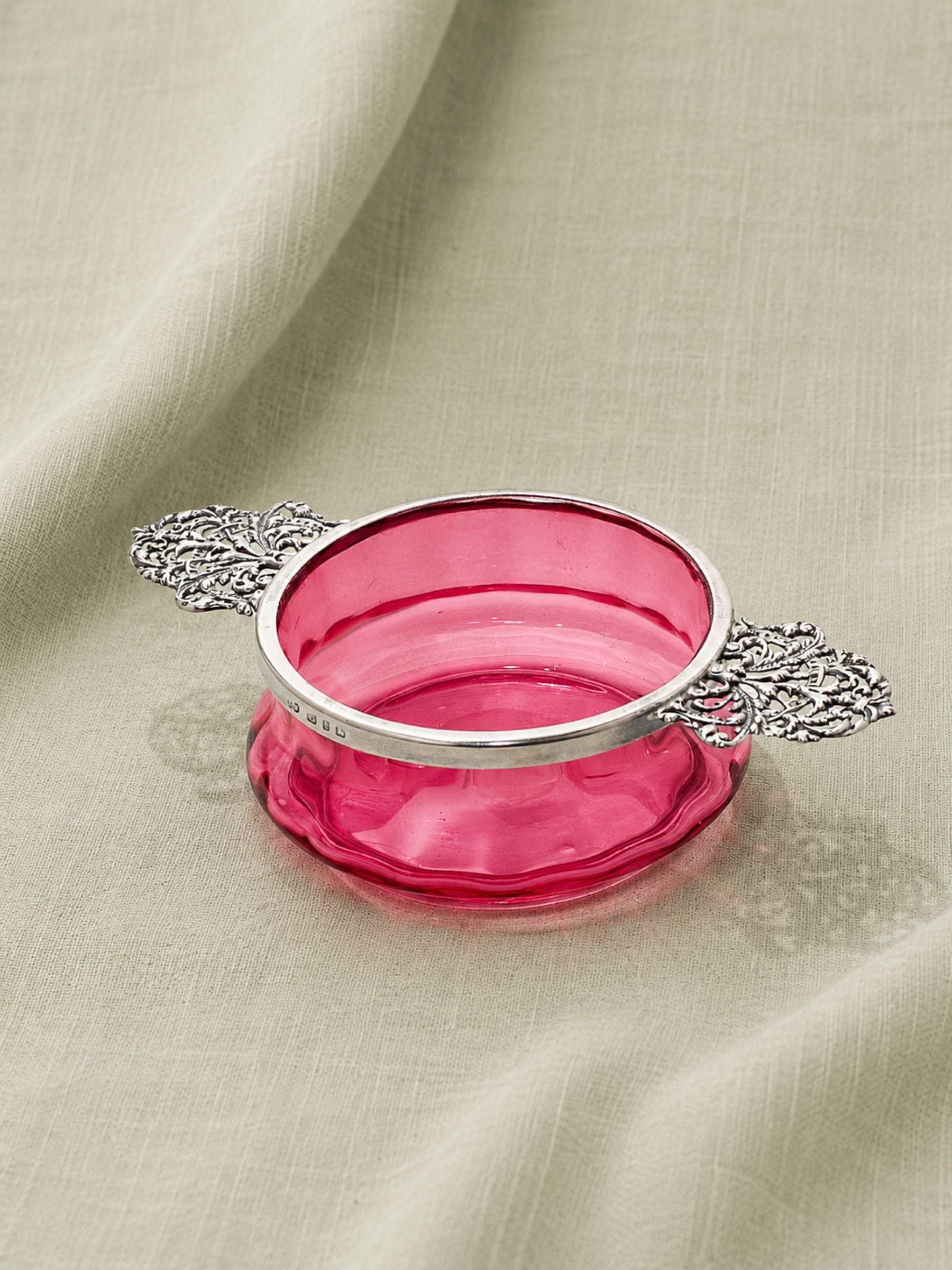 A cranberry glass bowl with ornate sterling silver rim and handles, resting on draped sage fabric.