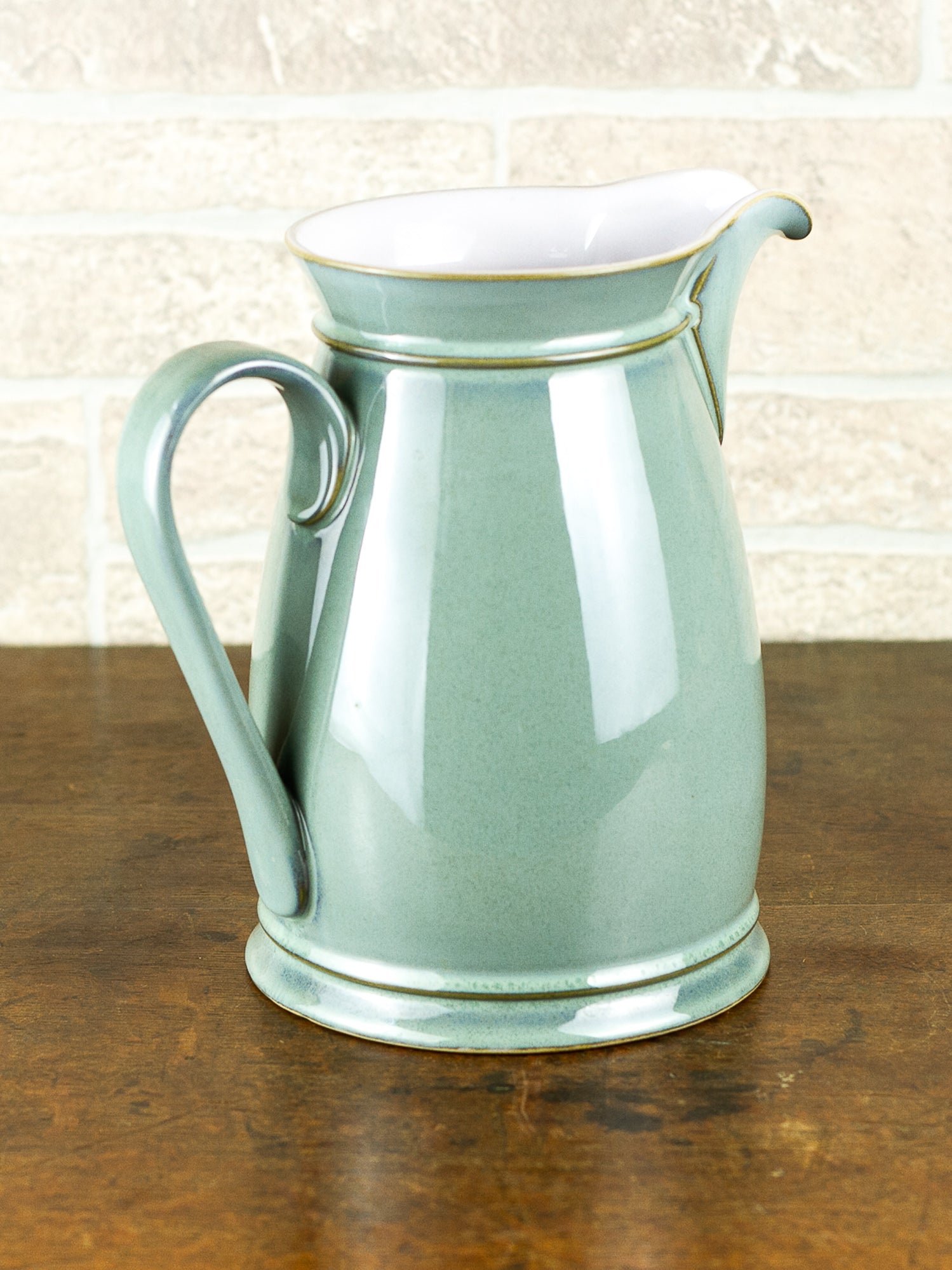 A glossy ceramic pitcher in a pale sage green glaze with a white interior, set on a dark wooden table against a light brick wall background.