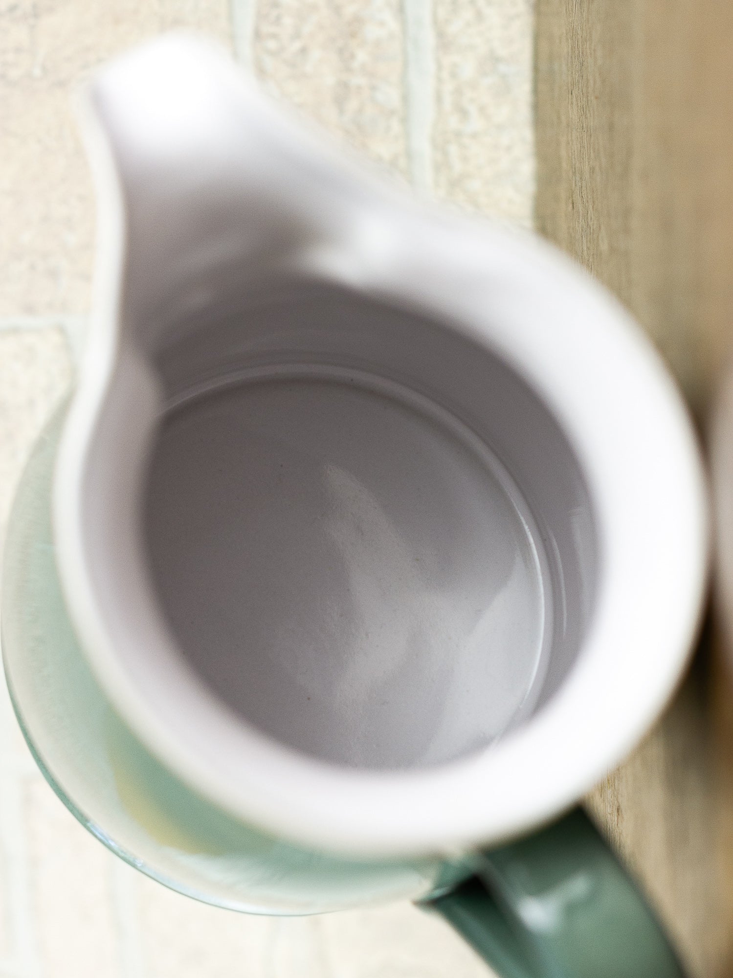 Close-up top-down view of a ceramic pitcher with a white interior and a pale green exterior, resting on a light wood surface.