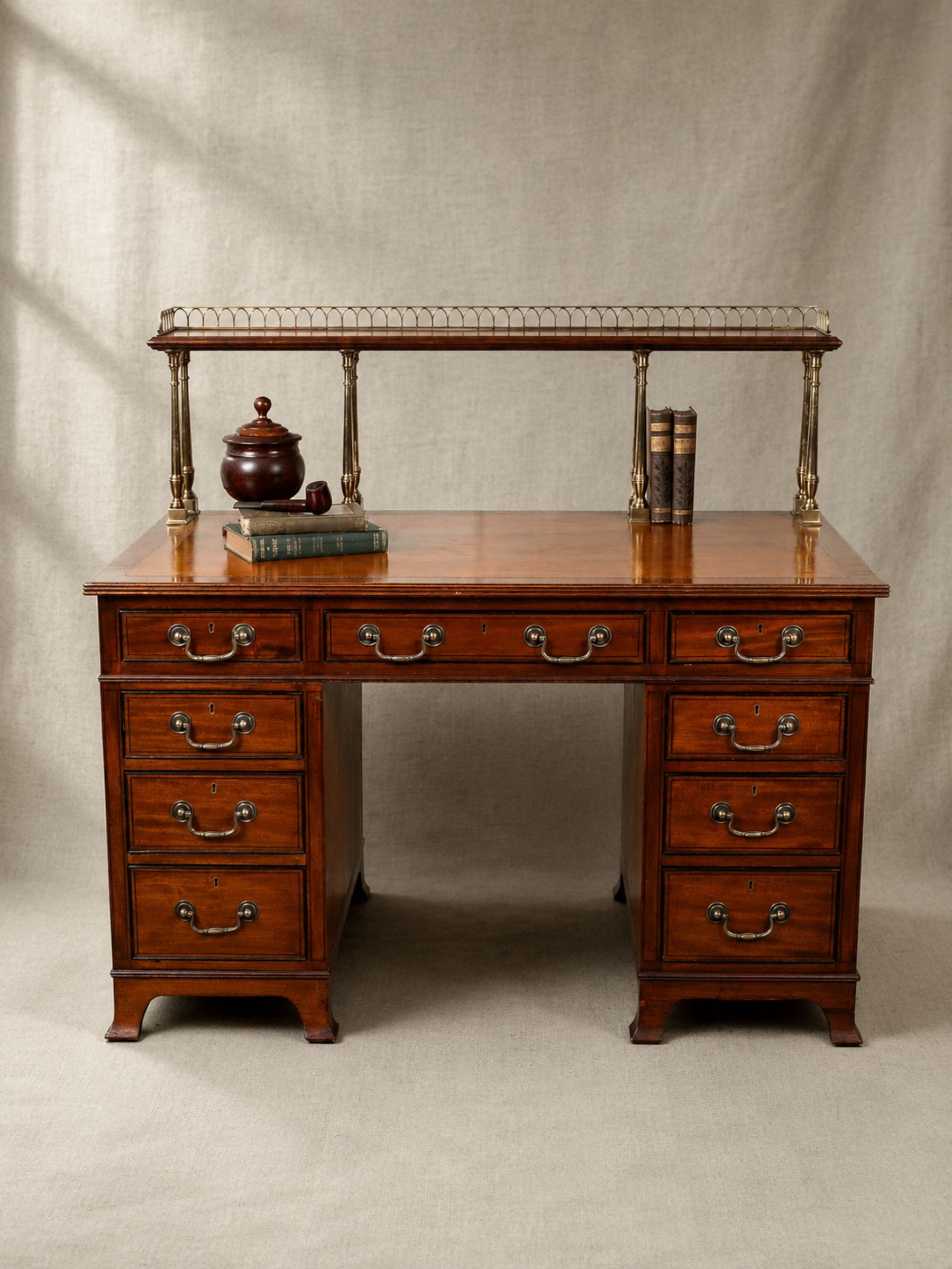Antique mahogany pedestal desk with brass gallery, multiple drawers and period books and pipe, photographed against a soft sage linen backdrop.