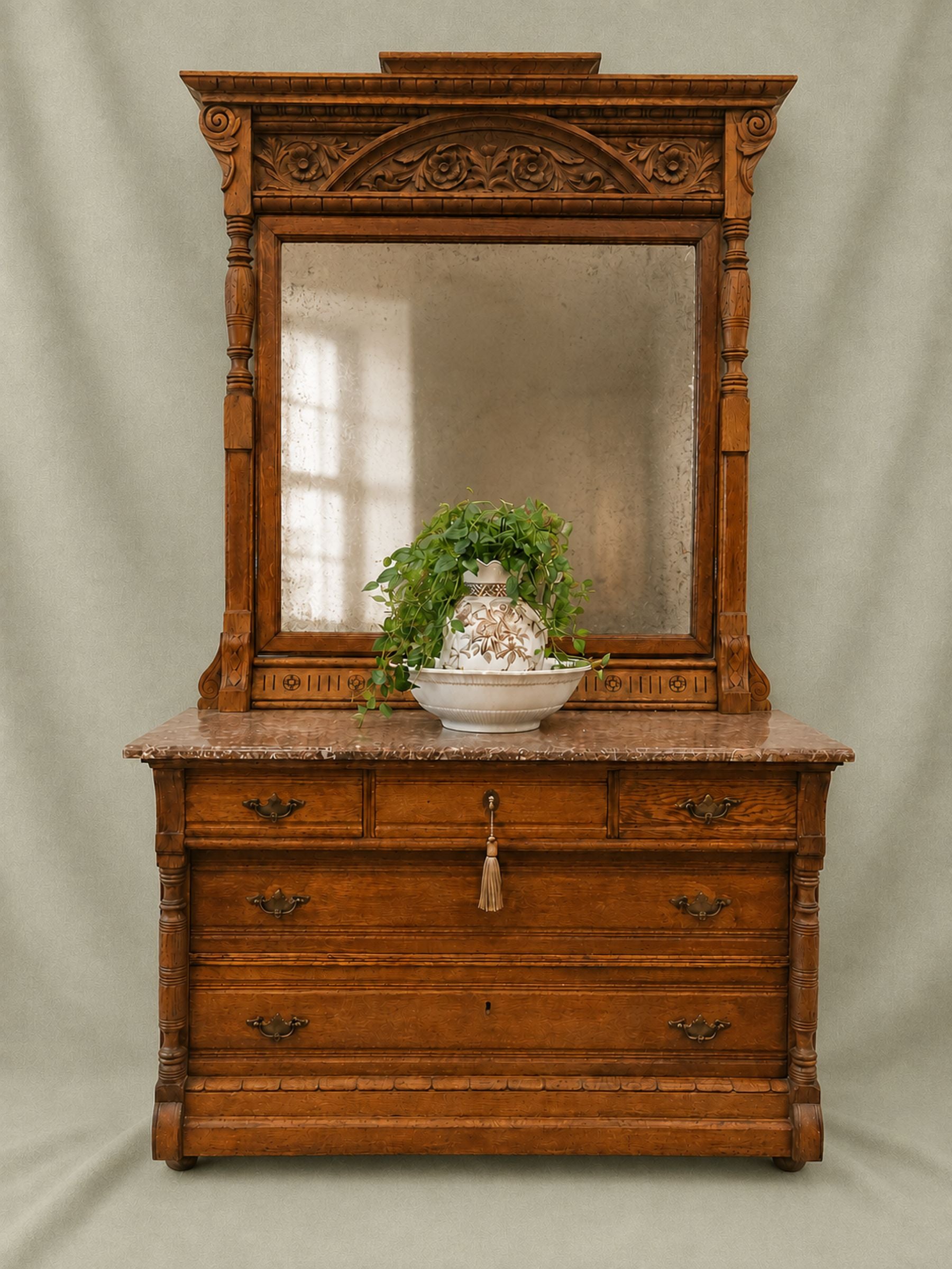 An antique carved wooden dresser with an attached mirror, featuring a marble top and a decorative ceramic vase with green foliage on top, set against a neutral draped backdrop.