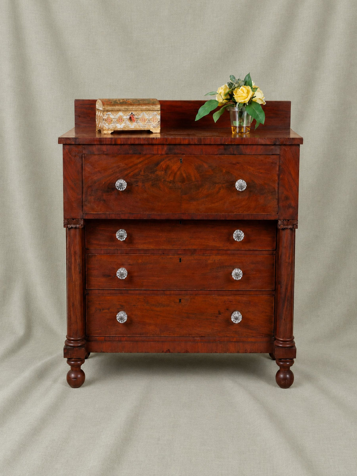 A antique mahogany chest of drawers with five drawers and round clear knobs, styled with a small wooden box and yellow flowers on top, set against a neutral draped fabric backdrop.