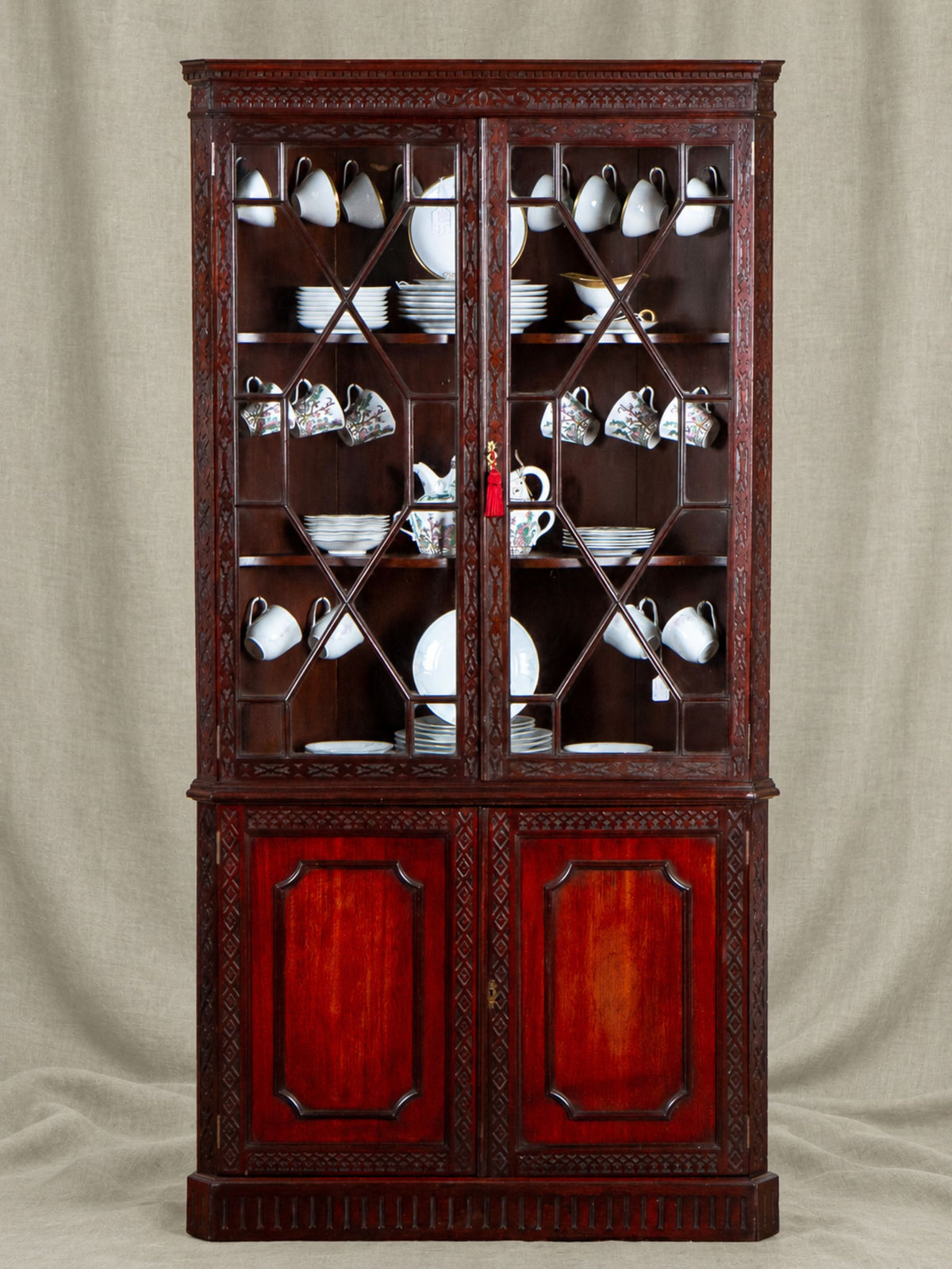 A tall dark reddish-brown wooden china cabinet with glass doors, filled with white teacups and plates, set against a neutral beige fabric backdrop.