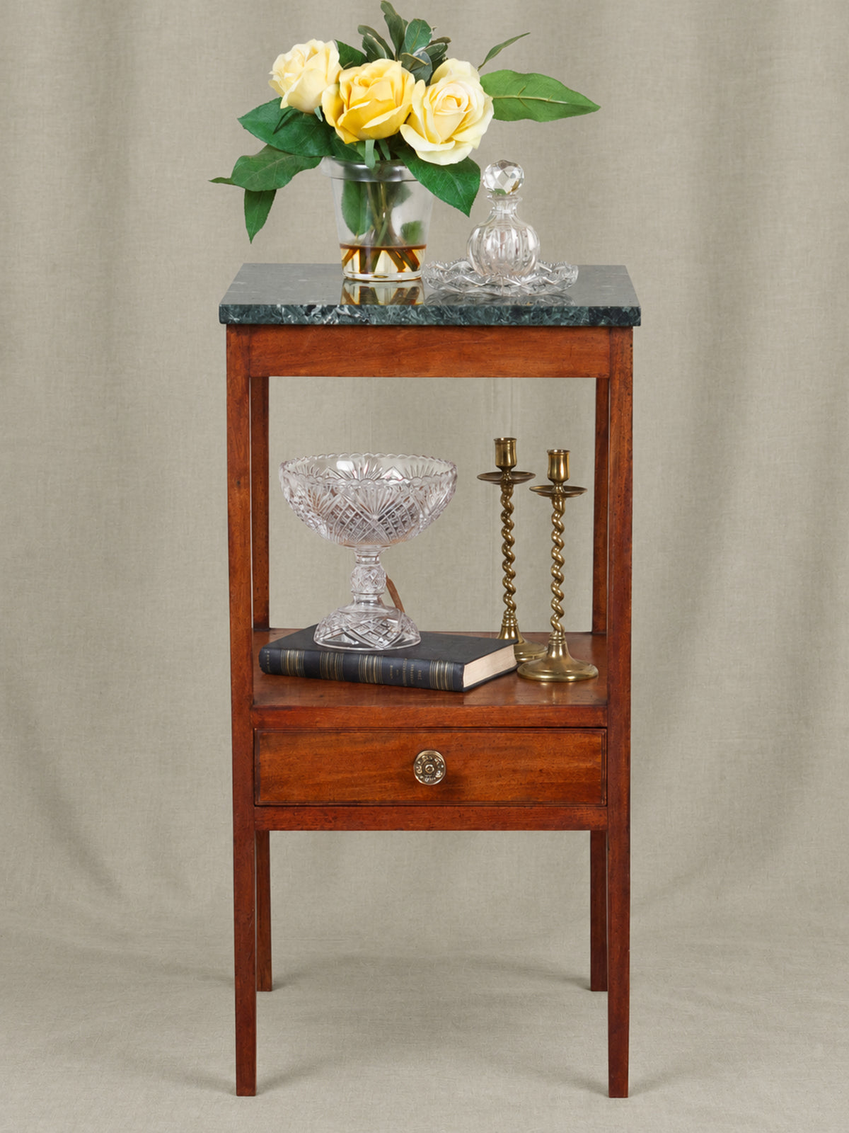 A wooden side table with a dark green marble top, featuring a single drawer and an open shelf styled with yellow roses, a glass bowl, candlesticks, and a book against a neutral beige backdrop.