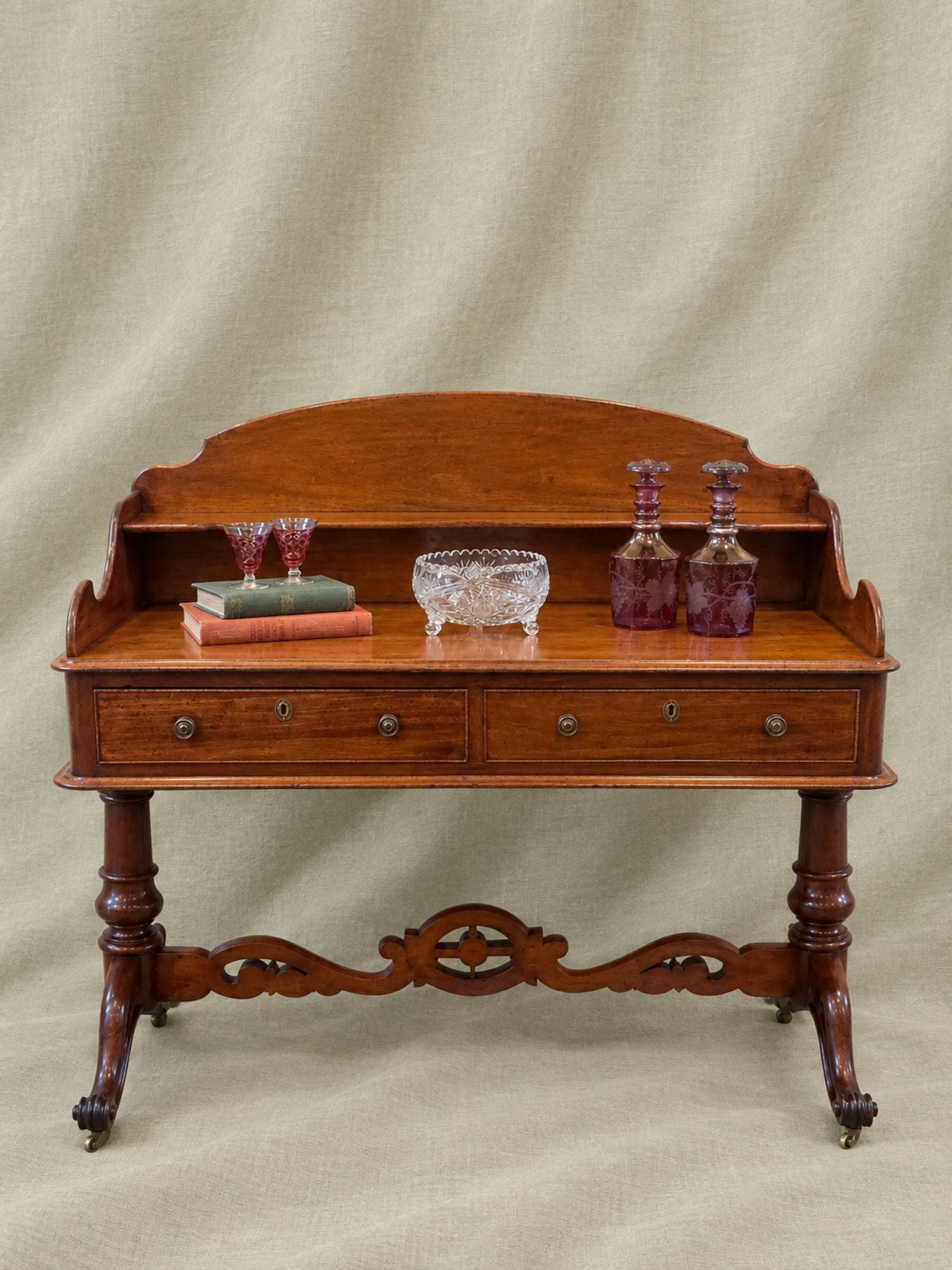 An antique wooden writing desk with a curved backrest, two drawers, and ornate turned legs, styled with books, a glass bowl, and decant bottles against a draped beige fabric backdrop.