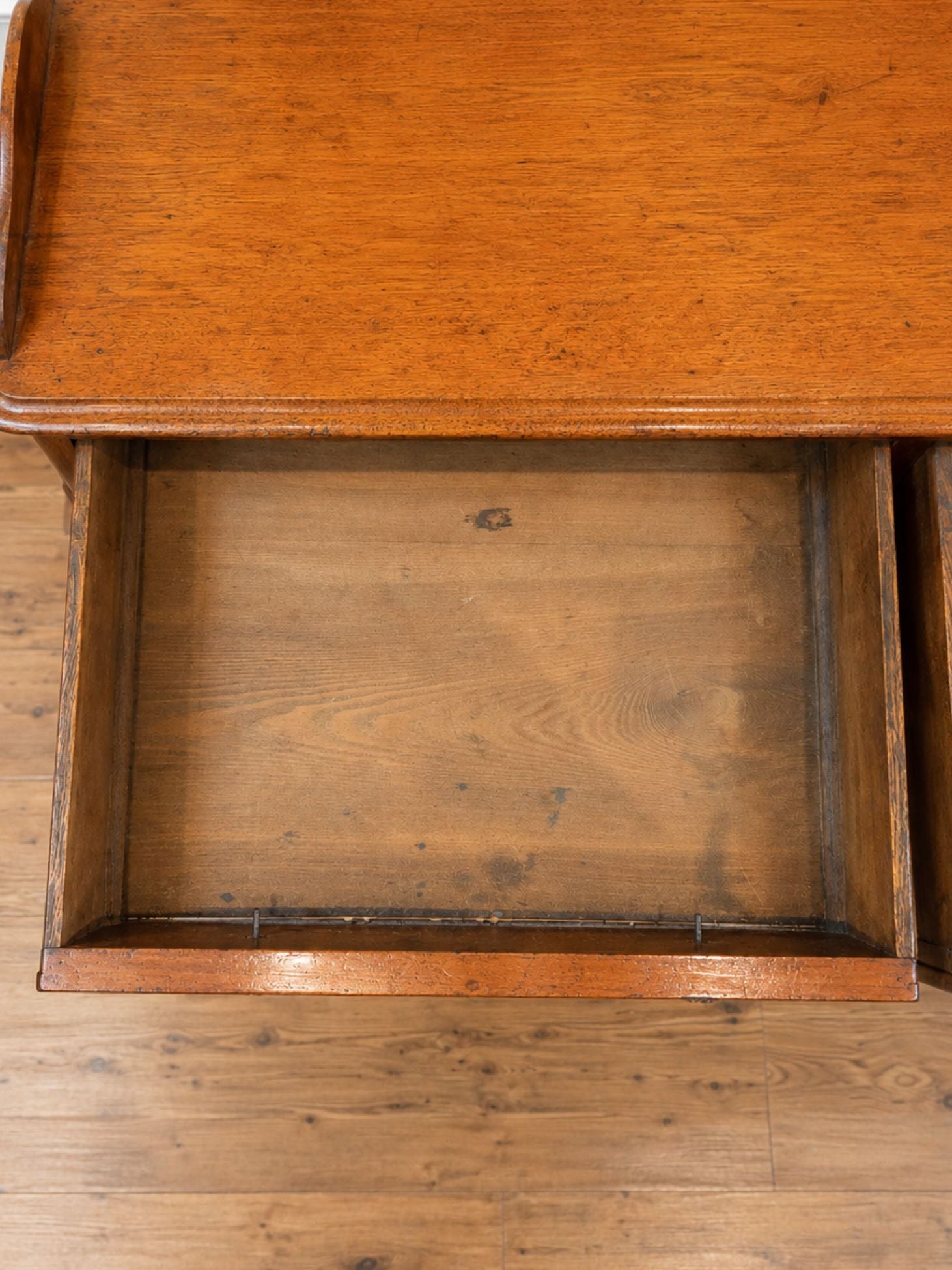 A close-up of a deskr bench with an open drawer, showing the warm brown wood grain and interior, set on a hardwood floor against a white baseboard.