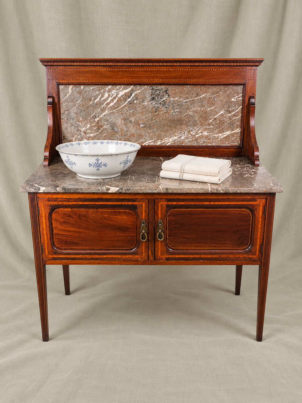 English mahogany inlaid washstand with  marble top, featuring two cabinet doors with brass ring pulls, The marble countertop is styled with a blue-and-white ceramic bowl and a folded cream linen cloth, all set against a neutral draped fabric backdrop.