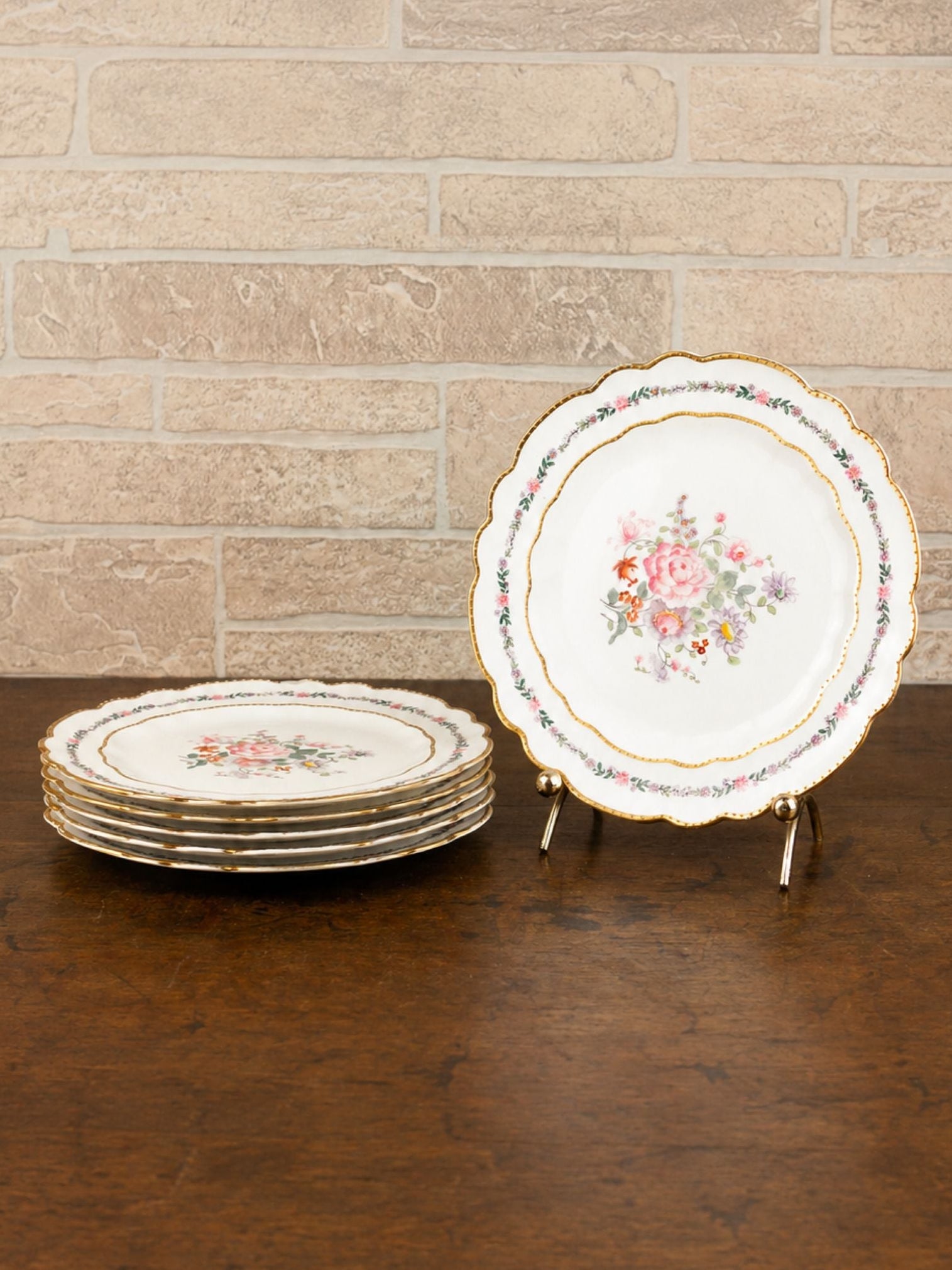 A set of vintage ceramic plates with floral designs, with one standing upright on a small display stand and a stack of five plates resting on a dark wooden table against a light brick wall.