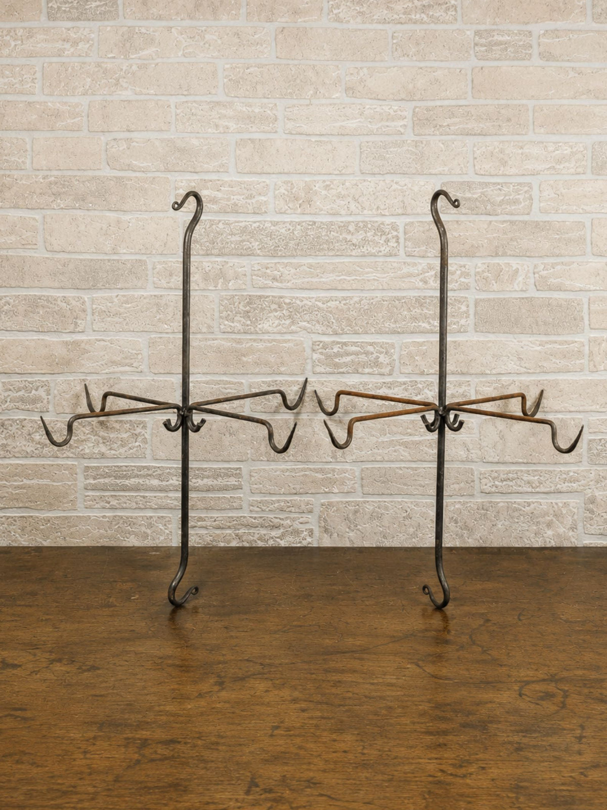 Two black metal meat hangers with multiple hooks, displayed on a dark wood floor against a light beige brick wall.
