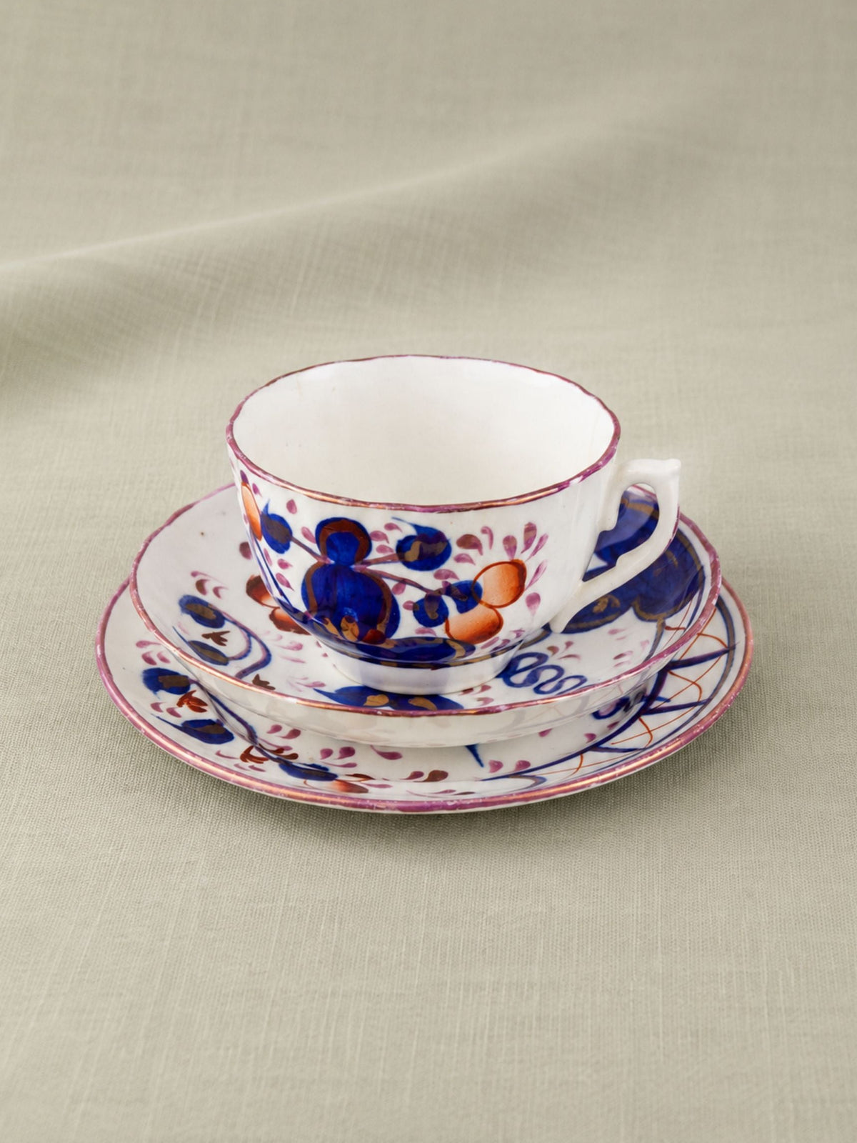 A white porcelain teacup and saucer set decorated with a hand-painted floral pattern in cobalt blue, orange, and red, set against a neutral textured fabric background.