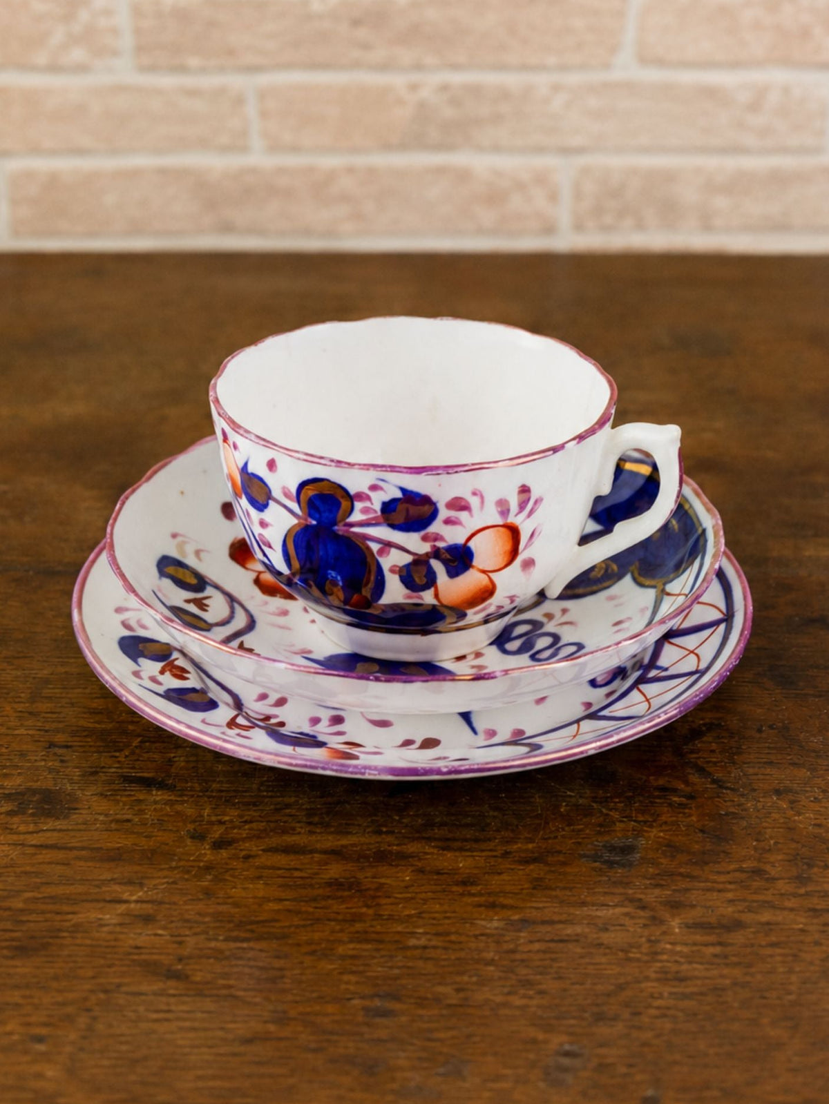 A white porcelain teacup and saucer set decorated with blue and orange floral patterns, resting on a dark wooden table against a light brick wall background.