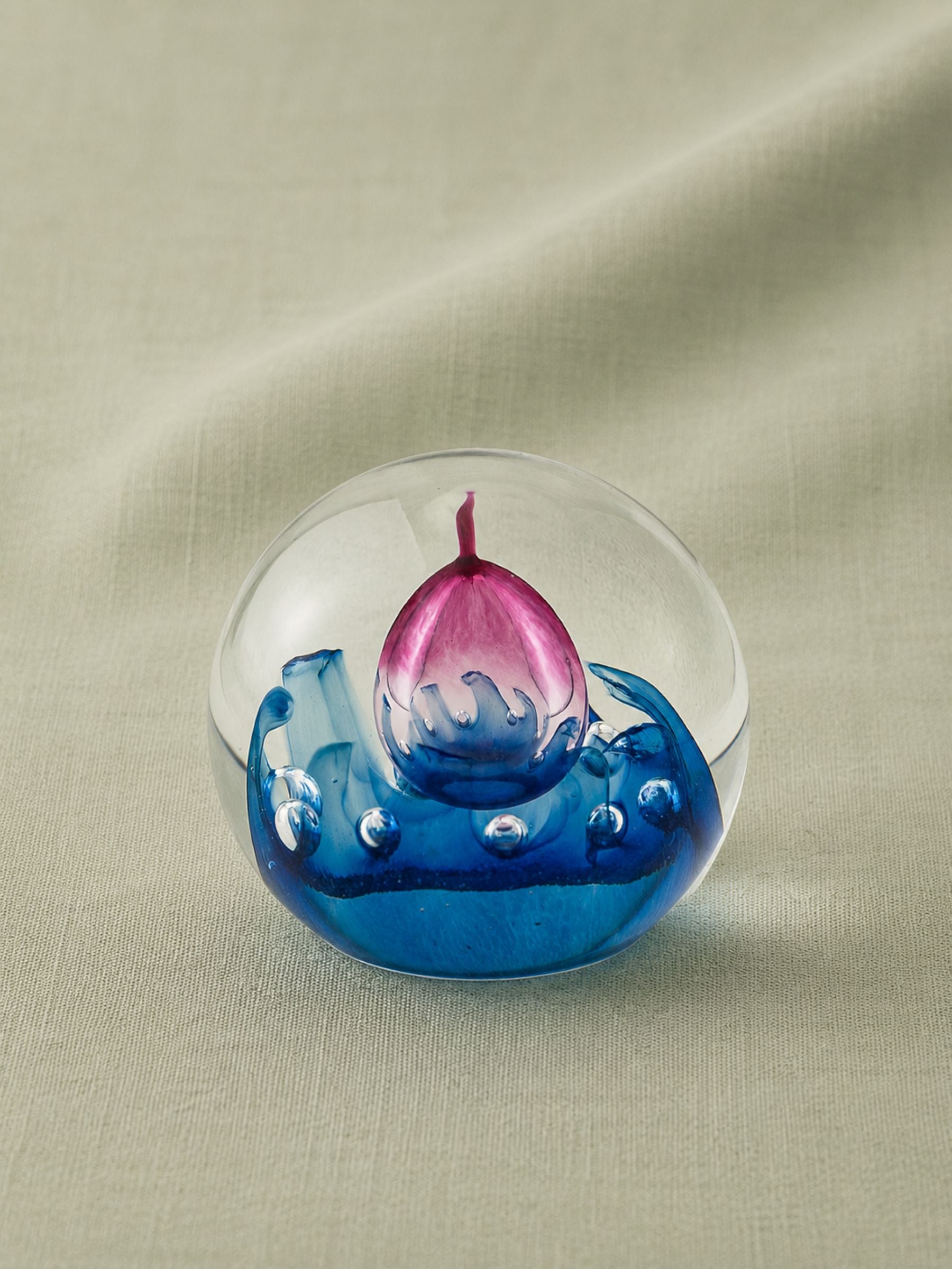 A clear glass paperweight featuring a blue and pink glass boat design, resting on a beige fabric background.