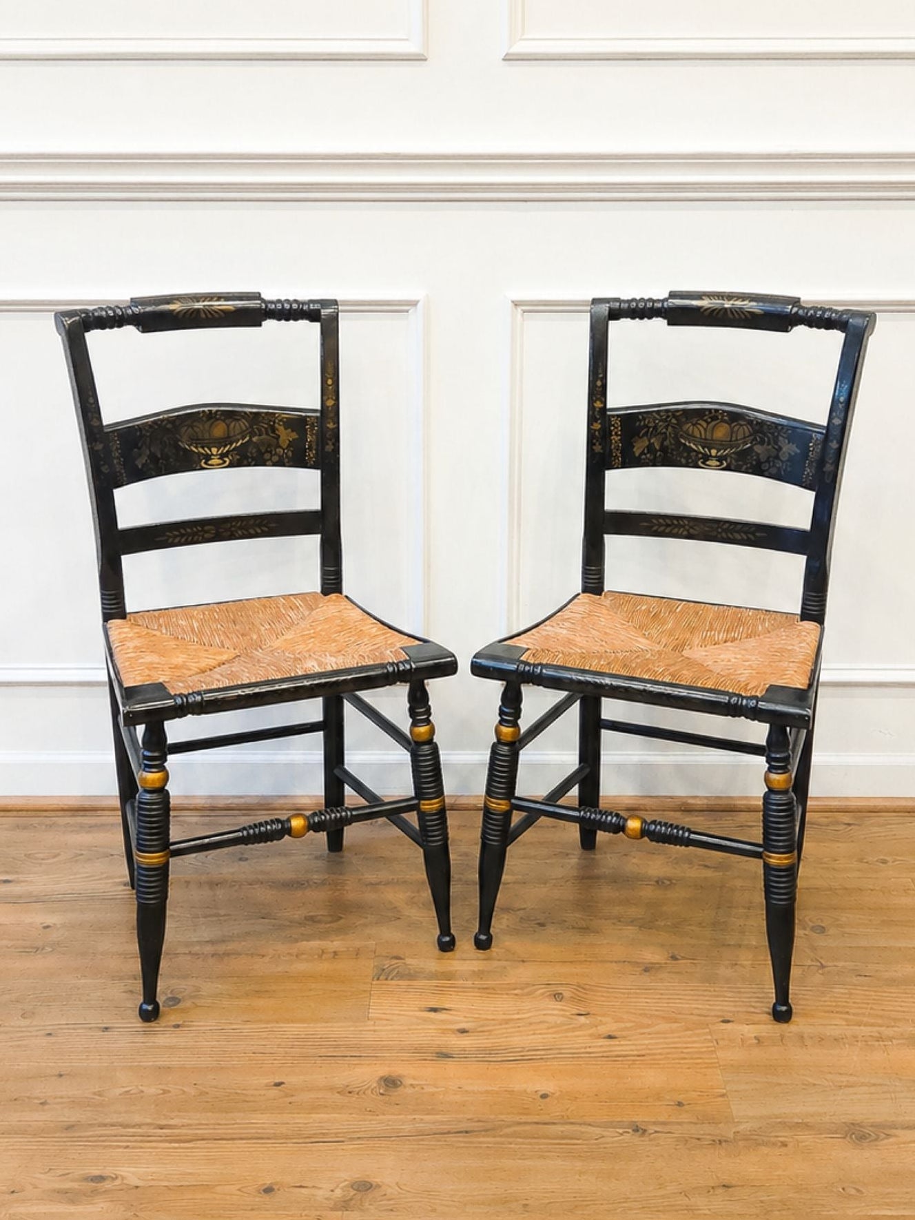 Two matching vintage Hitchcock black wooden ladder-back dining chairs with woven cane seats, positioned side-by-side on a light wood floor against a white paneled wall.
