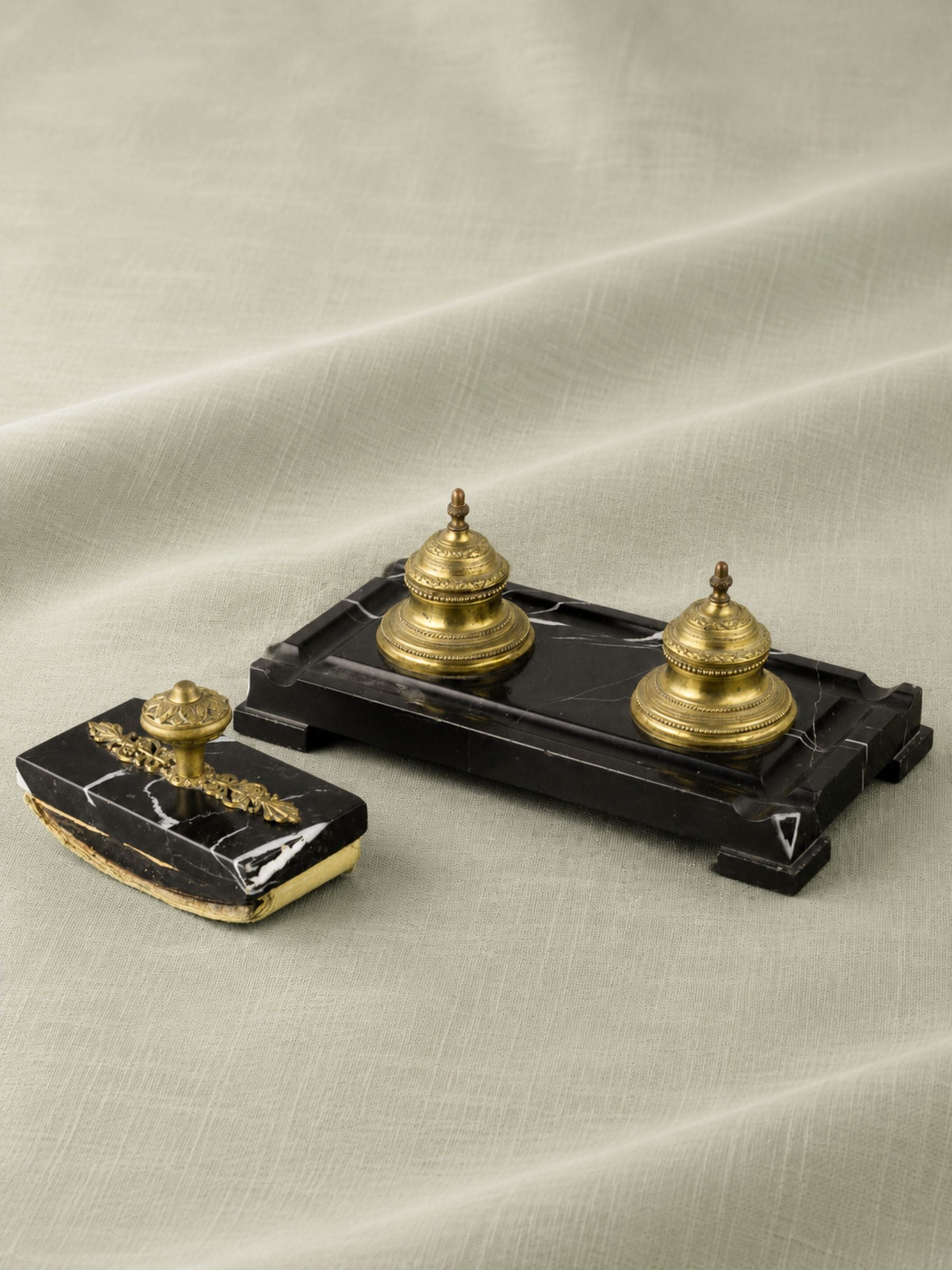A vintage-style double inkwell set on a draped light beige fabric background. The set includes a rectangular black marble base with two domed gold brass ink bottles, and a matching rectangular ink lid with gold decorative detailing on a black marble front.