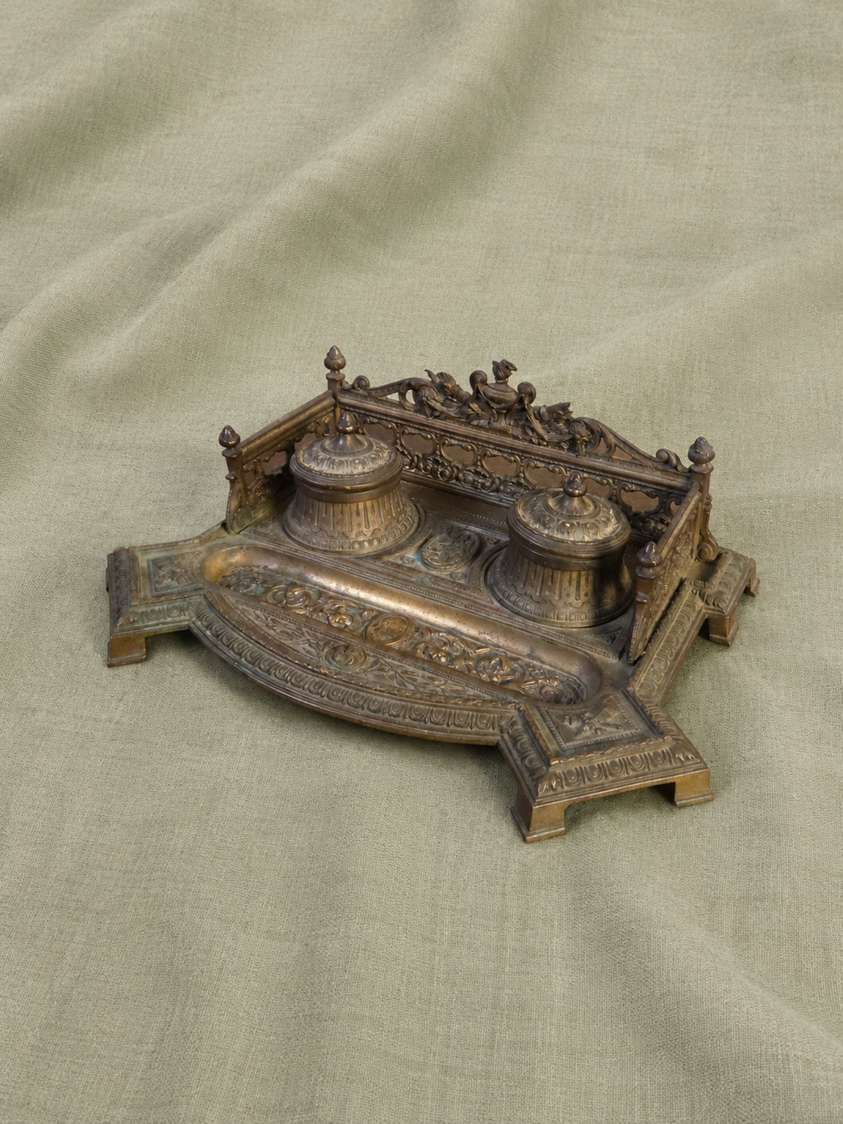 An ornate antique-style bronze metal Inkwell with two small domed vessels and carved decorative details, photographed on a draped beige fabric background.